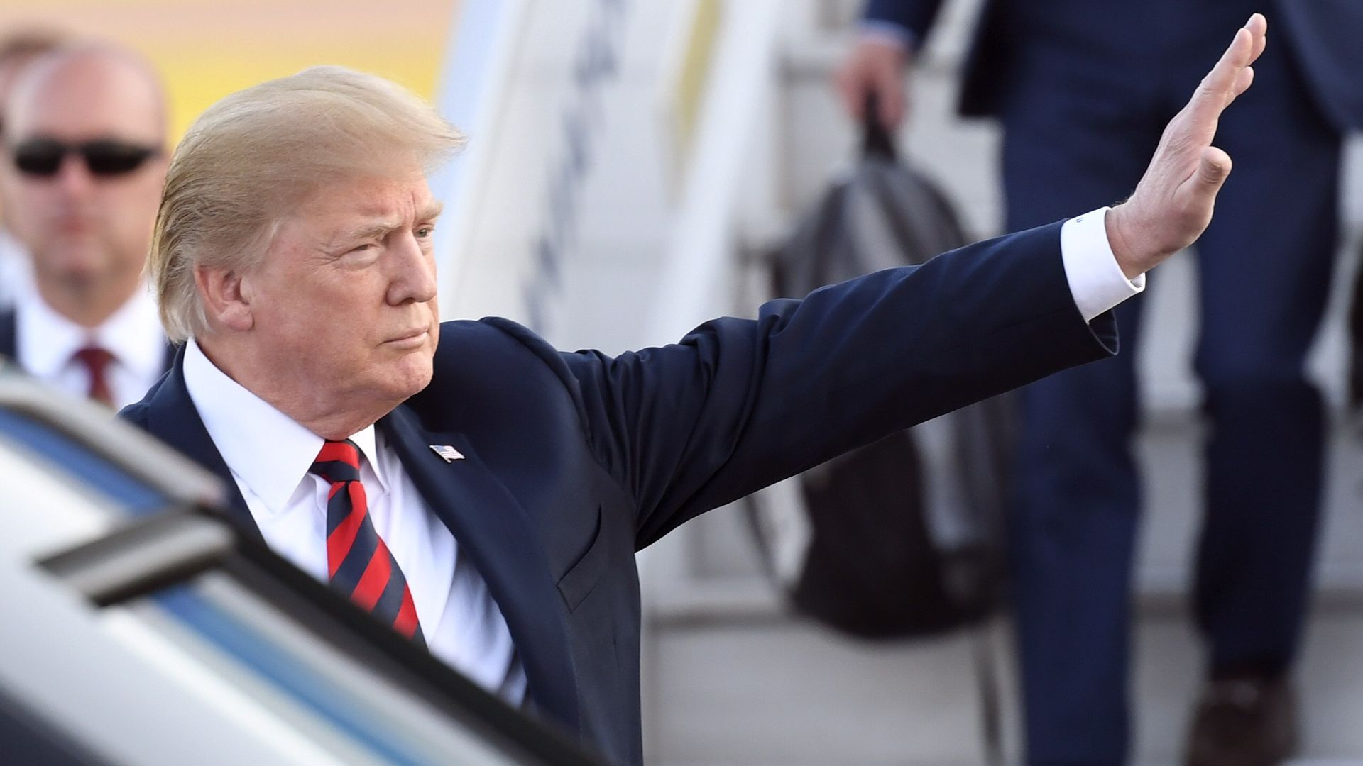 Donald Trump looks stern and raises his arm up with hand extended vertically as he enters car.