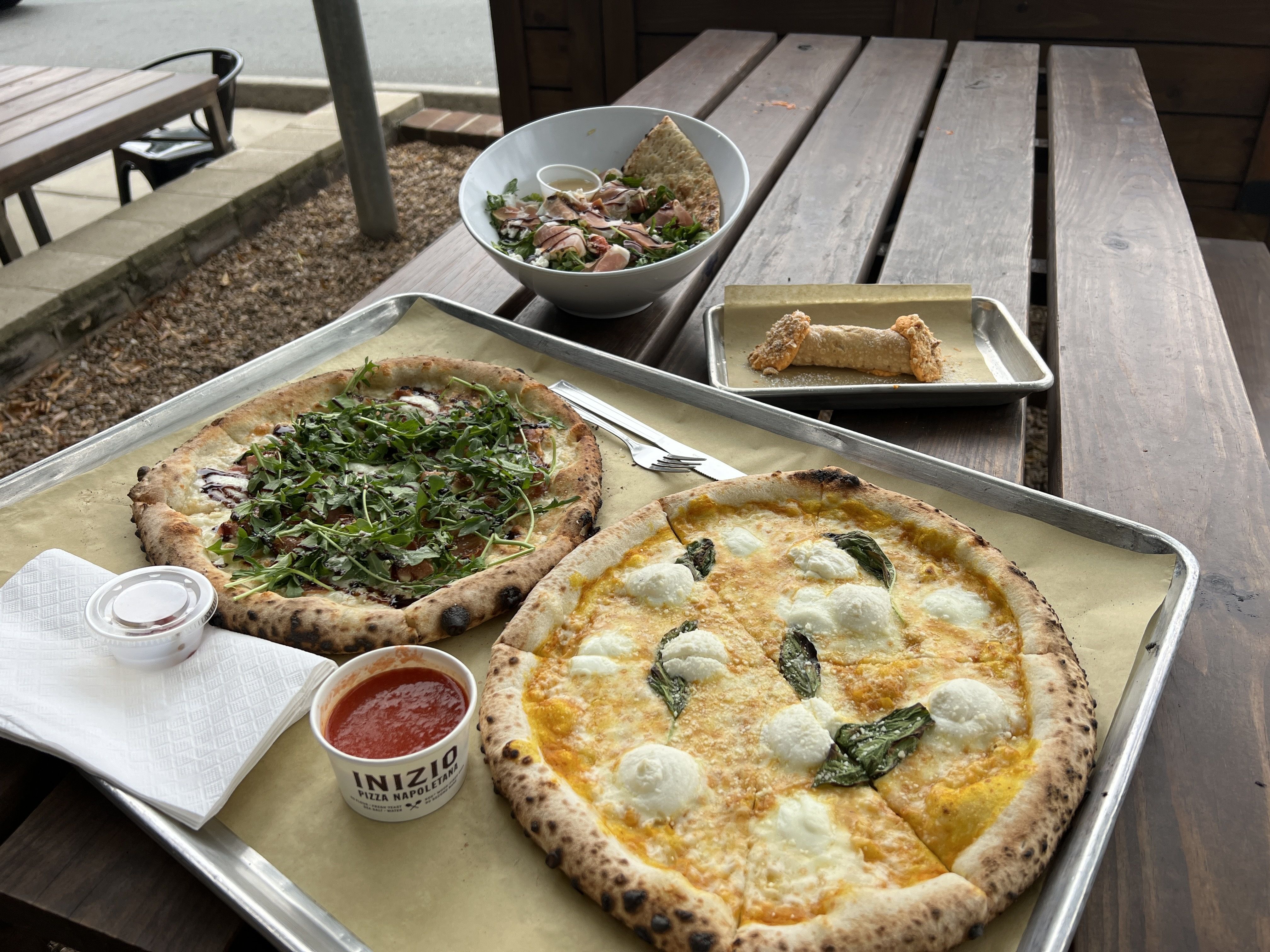 Wood-fired pizzas with fresh basil and arugula, a bowl of salad with prosciutto and pita chips, and a cannoli on a wooden picnic table outdoors.