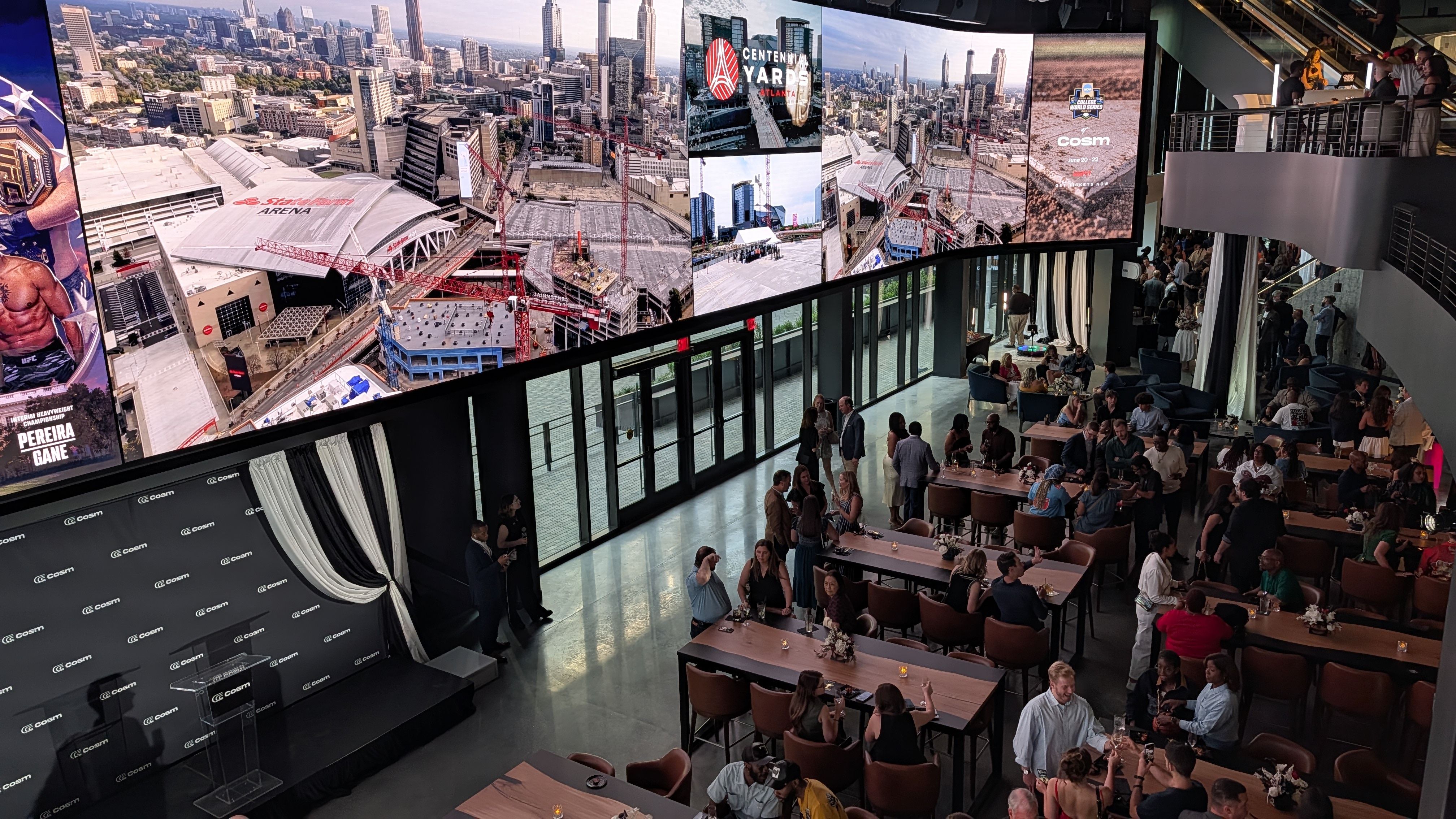 Interior of a modern event venue with a wall of screens showing the Atlanta skyline and red cranes, a stage on the left, and guests at long tables while others mingle on an upper balcony.
