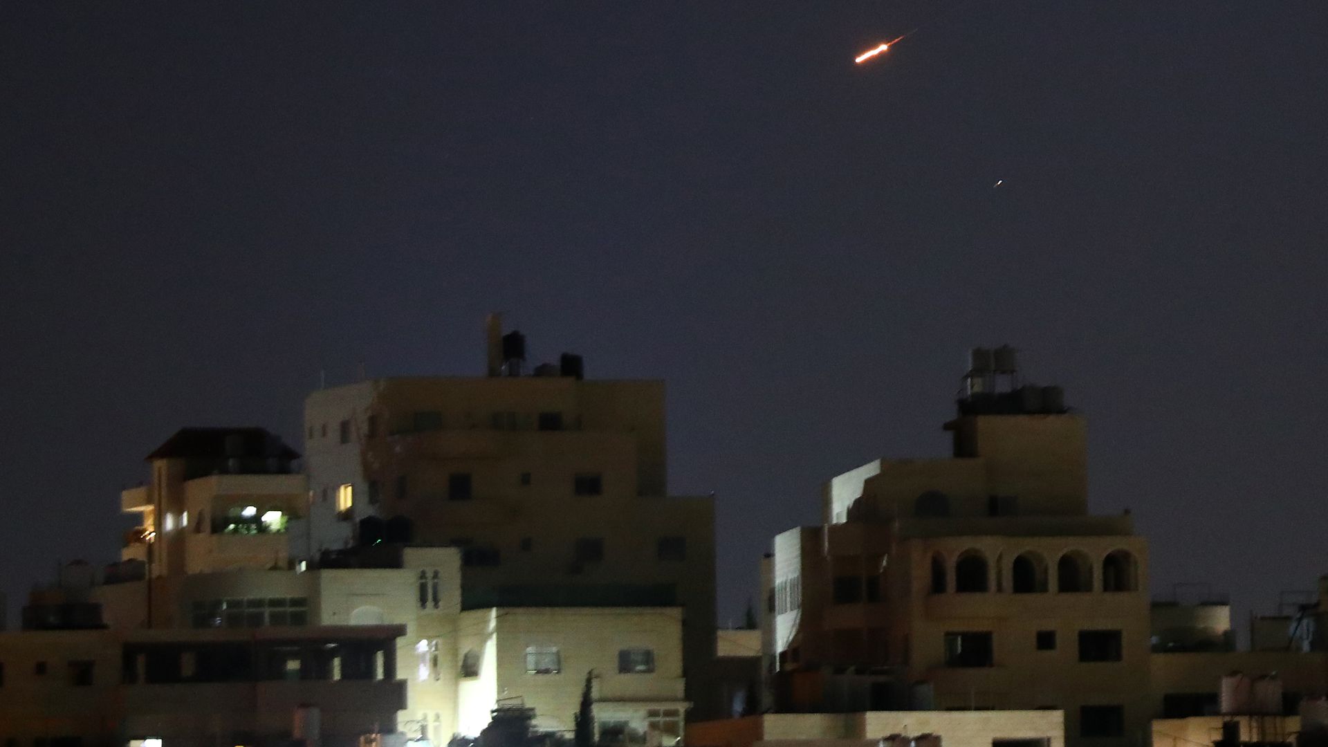 Nighttime cityscape with dark sky and lit windows along the bottom. A bright orange streak travels diagonally across the sky, with a small white streak nearby, like a meteor or rocket trail.