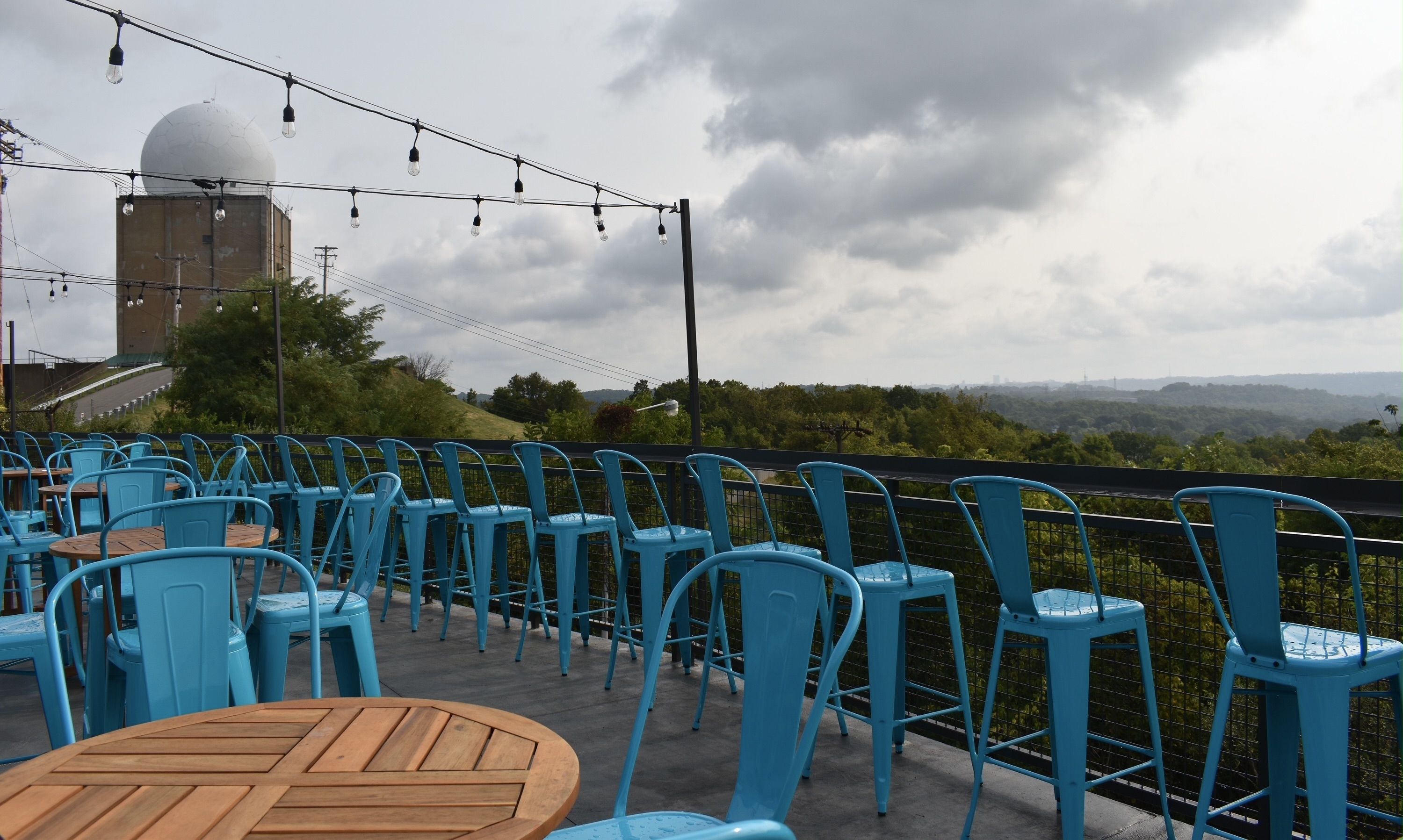 Outdoor rooftop with bright cyan metal chairs and round wooden tables, string lights overhead. A large white dome sits on a building to the left, under a cloudy sky and distant hills.