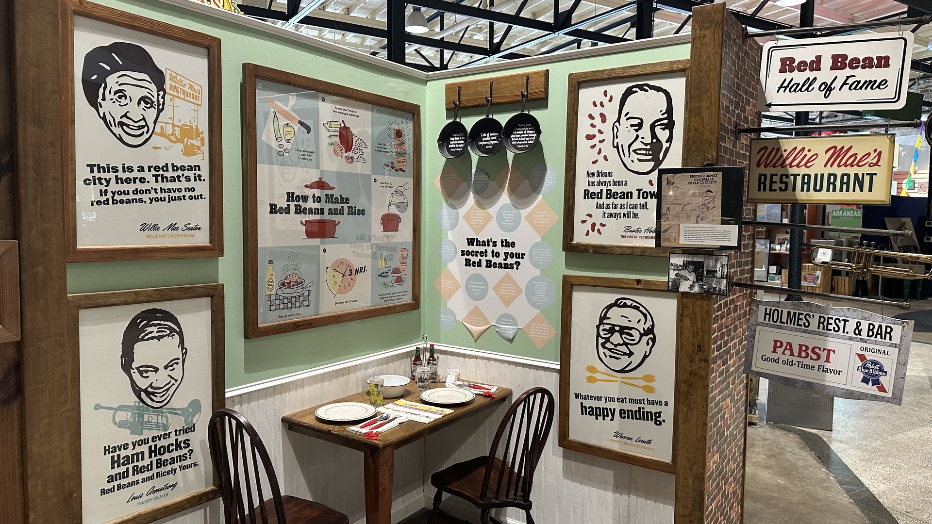 Corner of a retro restaurant display with wood-framed posters about red beans and rice, portraits, and signs like "Red Bean Hall of Fame" and "Willie Mae's Restaurant" on green walls.