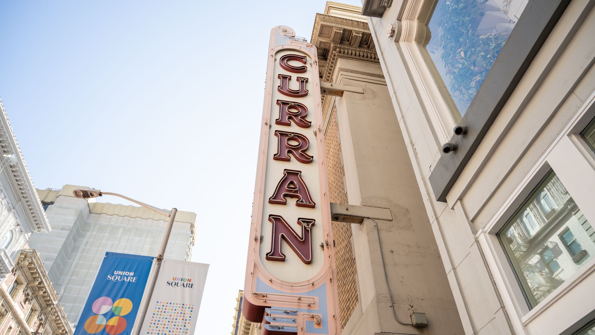 Urban street view with a tall pink vertical neon sign showing red letters on a beige building, blue sky, glass storefronts, and Union Square banners along the street.