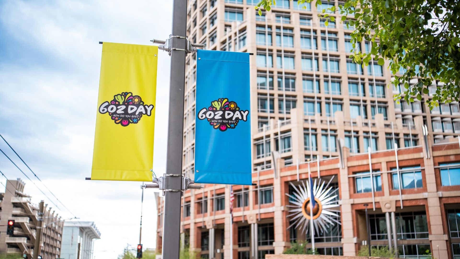 Two vertical banners, yellow and blue, hang from a lamppost with a colorful "602 DAY" logo. City buildings, a starburst sculpture, and green leaves frame the scene.