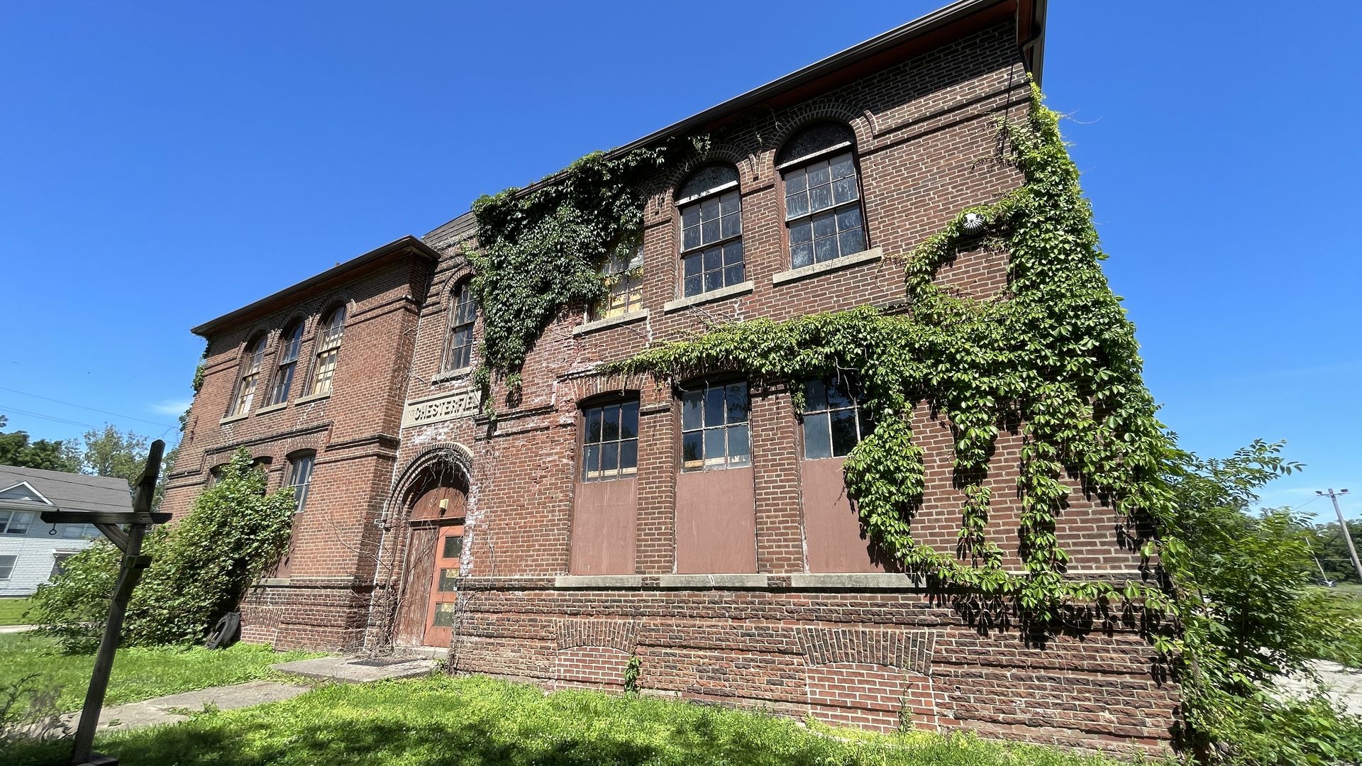 Old red brick building with arched windows, ivy creeping over its walls, bright blue sky, boarded lower windows; a neglected, ivy-covered structure.