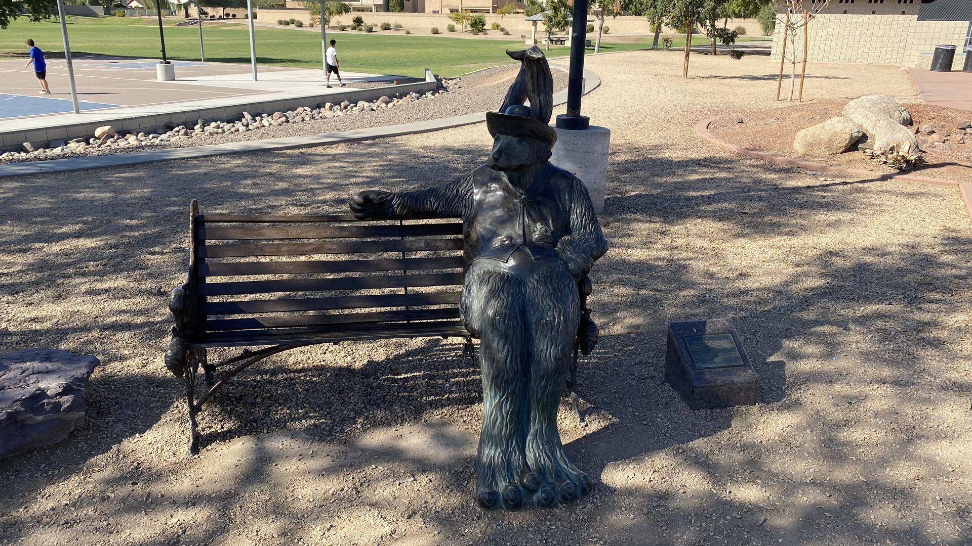 A metal sculpture of a dog with a human's body wearing a hat and sitting on a bench. 