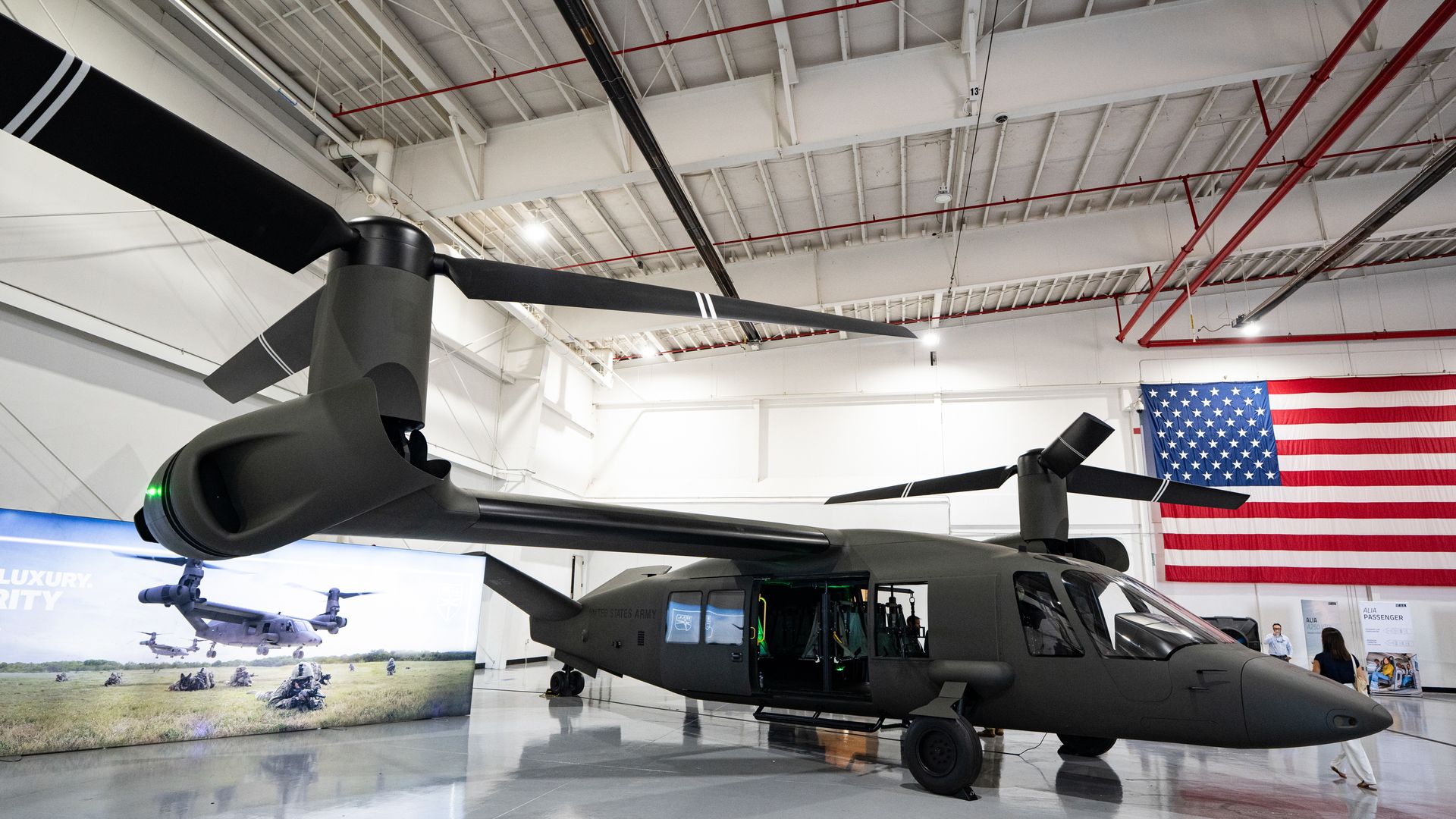 A large, black-in-color tiltrotor aircraft is seen inside a bright room. A large American flag hangs on the wall.