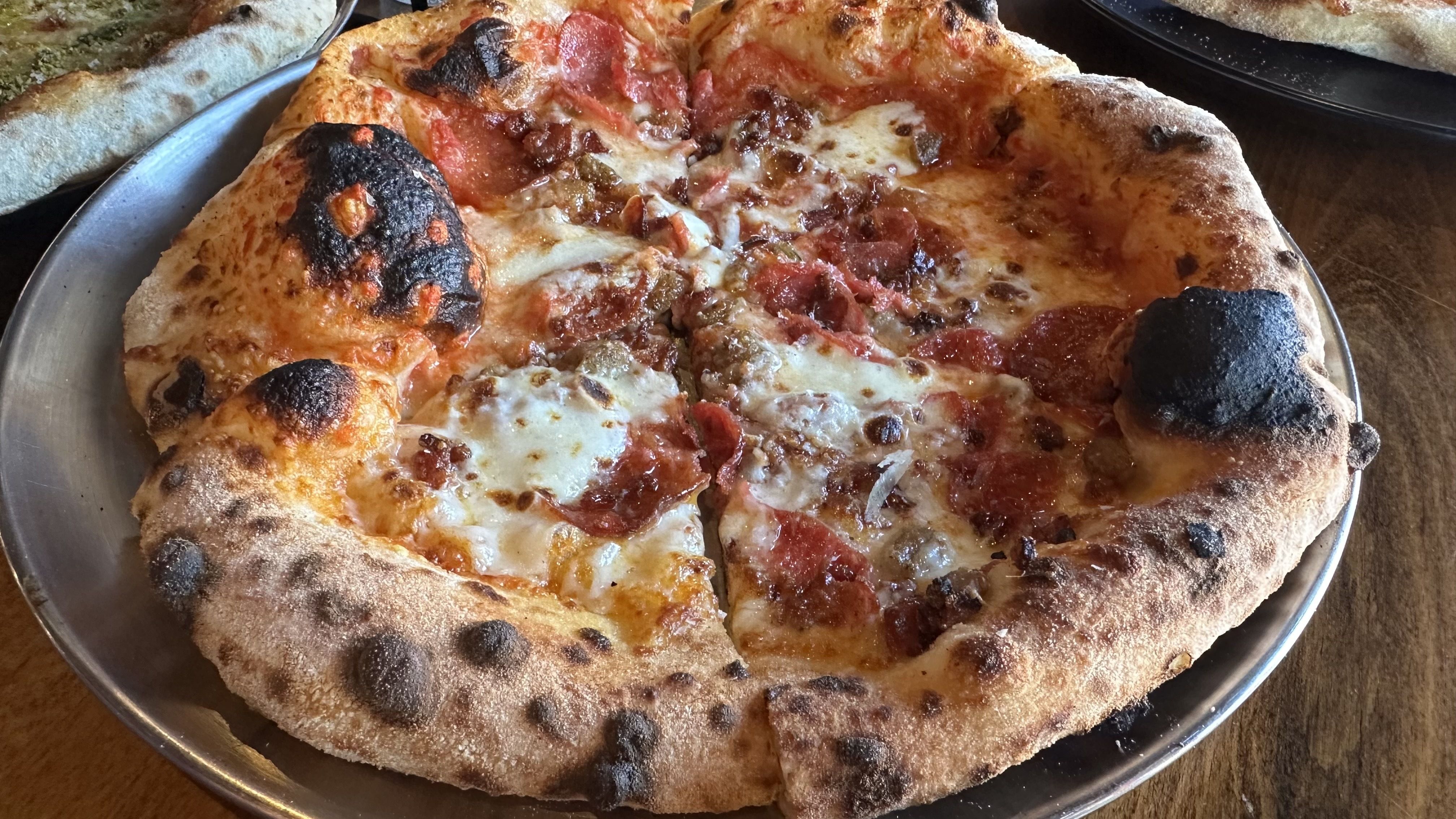 Close-up of a large round pepperoni and bacon pizza on a metal tray; bubbly cheese, browned edges, and dark, charred spots on a thick, blistered crust.