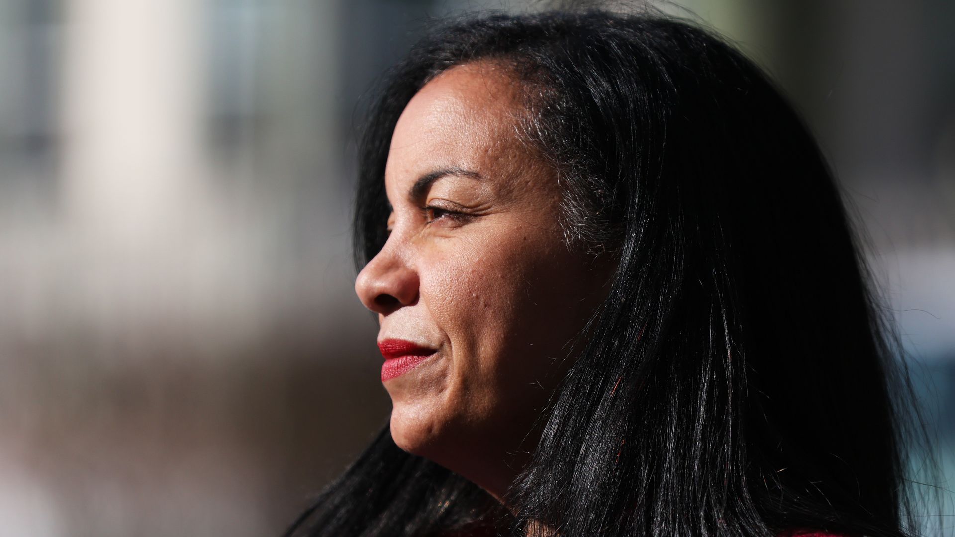 Portrait of a woman with dark hair and red lipstick, wearing a red coat, looking thoughtfully to the left against a blurred outdoor background.
