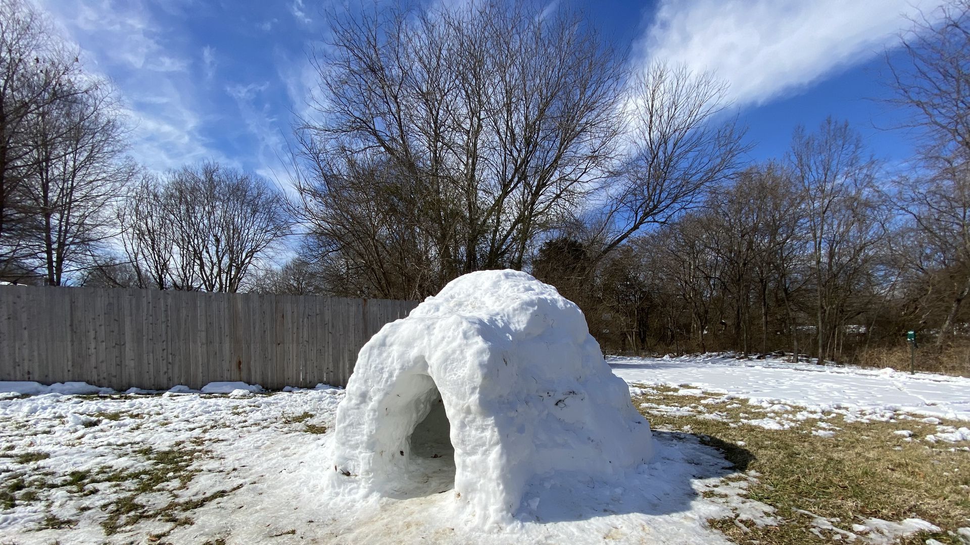 An igloo sits on grass half hidden by snow