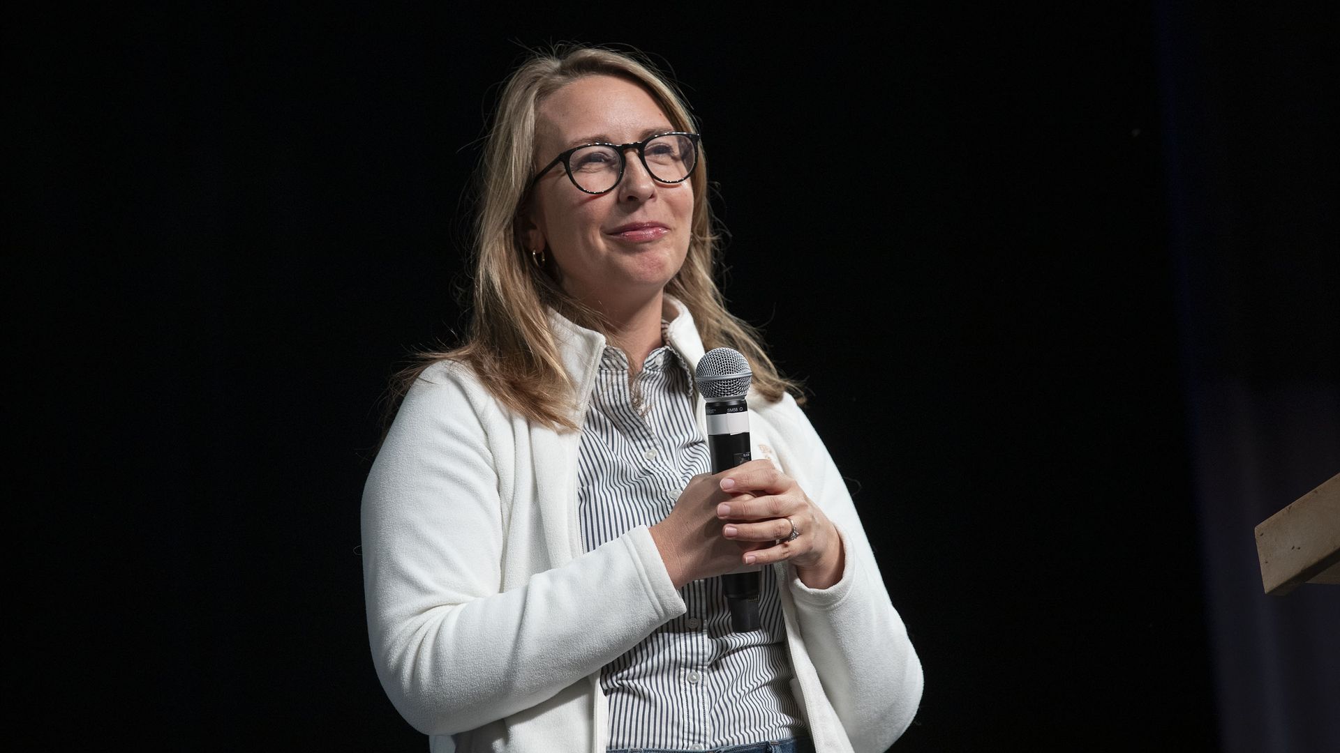 Rep. Hillary Scholten, wearing a white fleece jacket over a white-and-black button down, holds a microphone in both hands in front of a black backdrop.