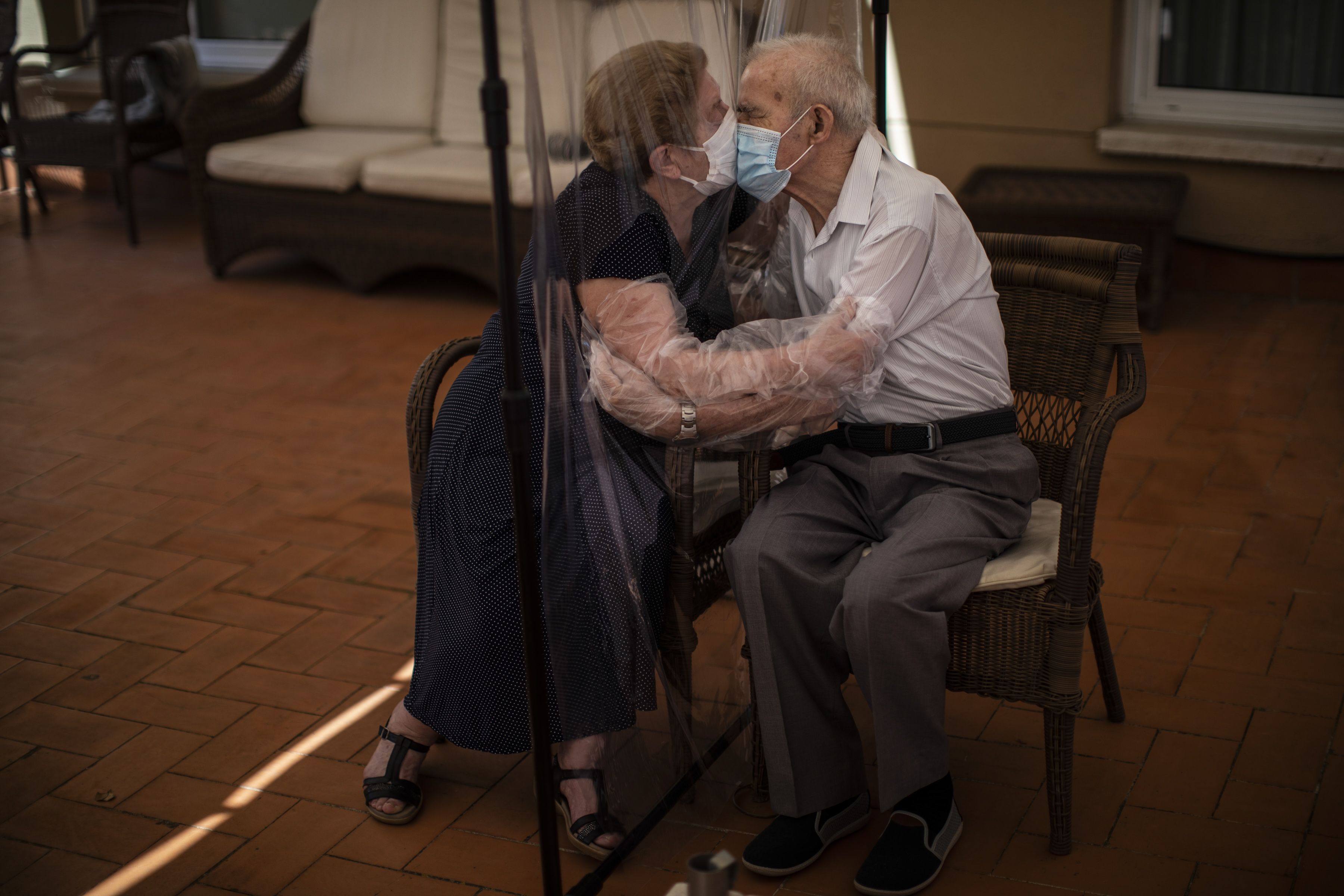 Elderly couple wearing face masks share a kiss through a clear plastic shield on a sunlit patio, seated on wicker chairs with a tiled floor.