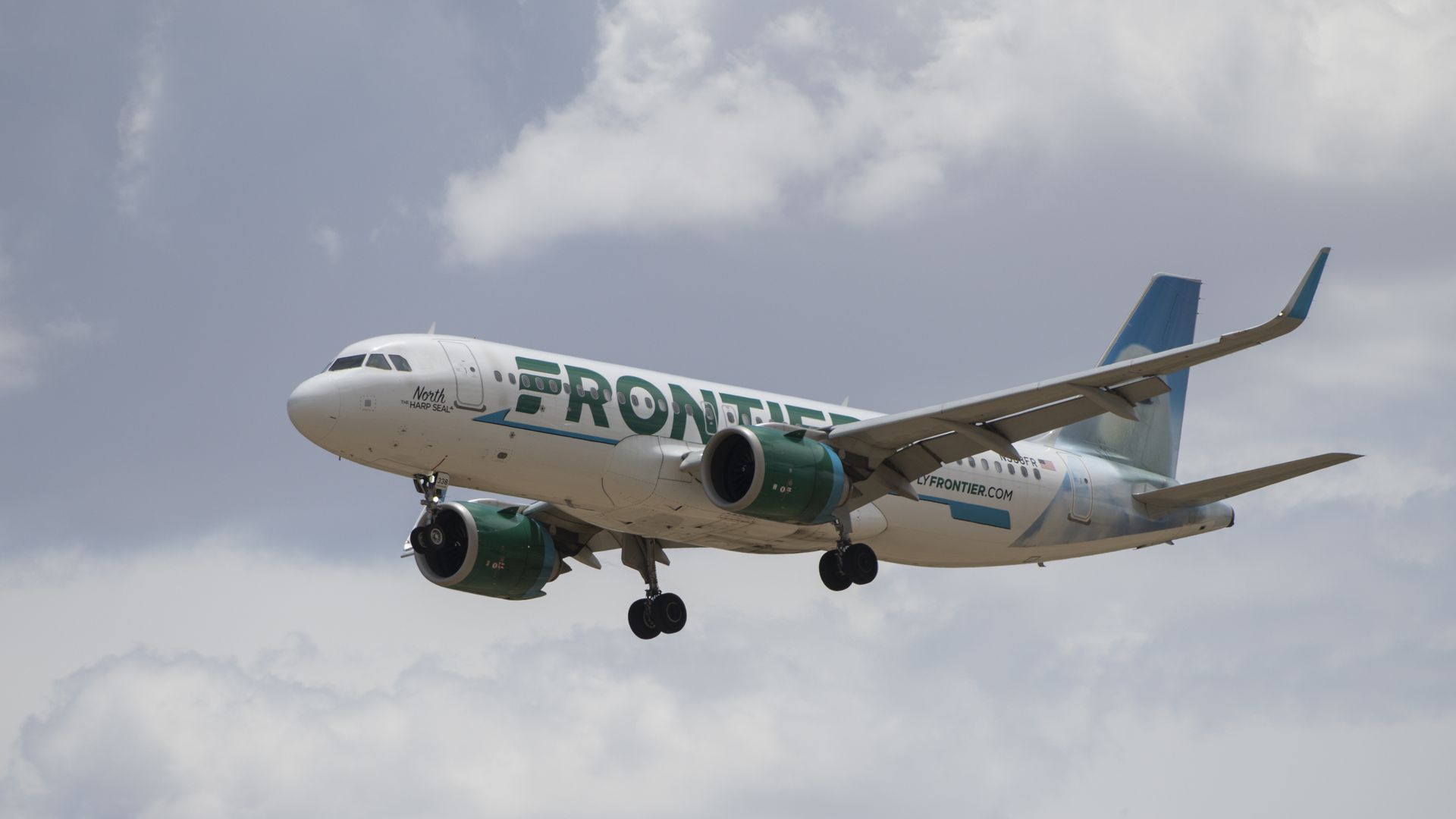 Frontier Airlines jet in flight, nose left, landing gear down, white fuselage with green engine and large FRONTIER lettering, blue tail, against a cloudy sky.