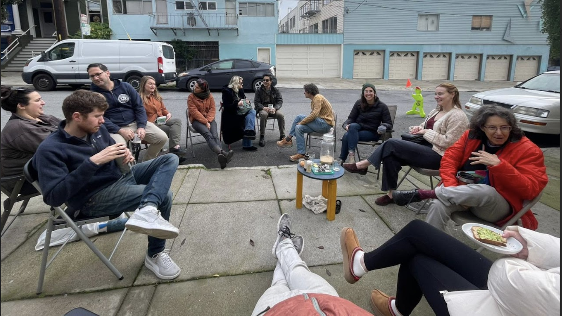 Group of twelve people sitting in a circle on folding chairs outside on a cloudy day, chatting and enjoying drinks, in front of blue and white residential buildings.