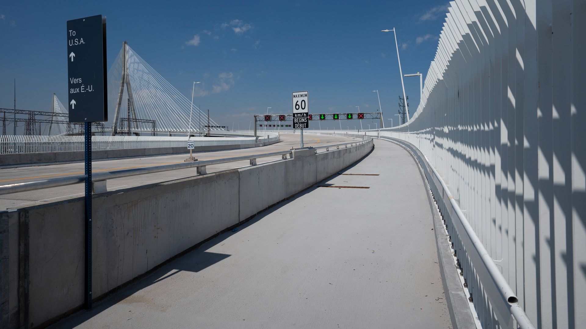 A pedestrian path on a bridge under a clear blue sky. A tall blue sign reads "To U.S.A." with an upward arrow. On the right, a white barrier casts shadows; distant cable-stayed bridge visible.
