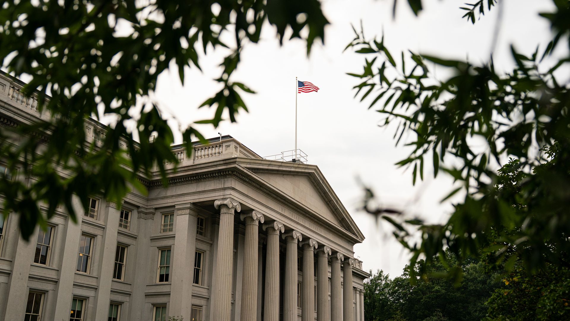 View of a classical white stone building with tall columns, framed by green tree branches. An American flag flies atop the building under a cloudy sky.