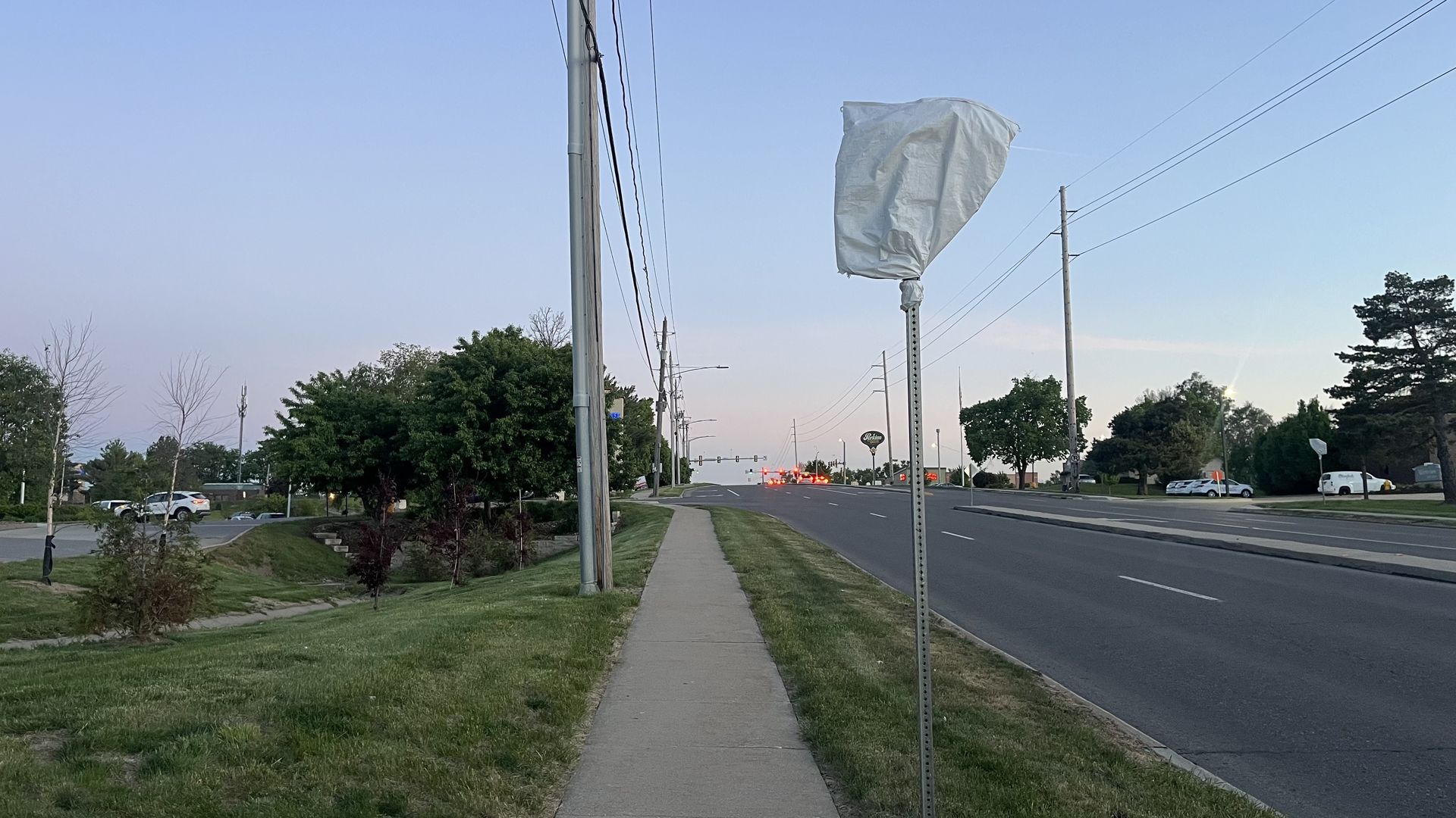 Wide suburban street scene with a sidewalk and grassy median. A large white bag covers a street sign on a pole; power lines, trees, and distant traffic under a clear blue sky.