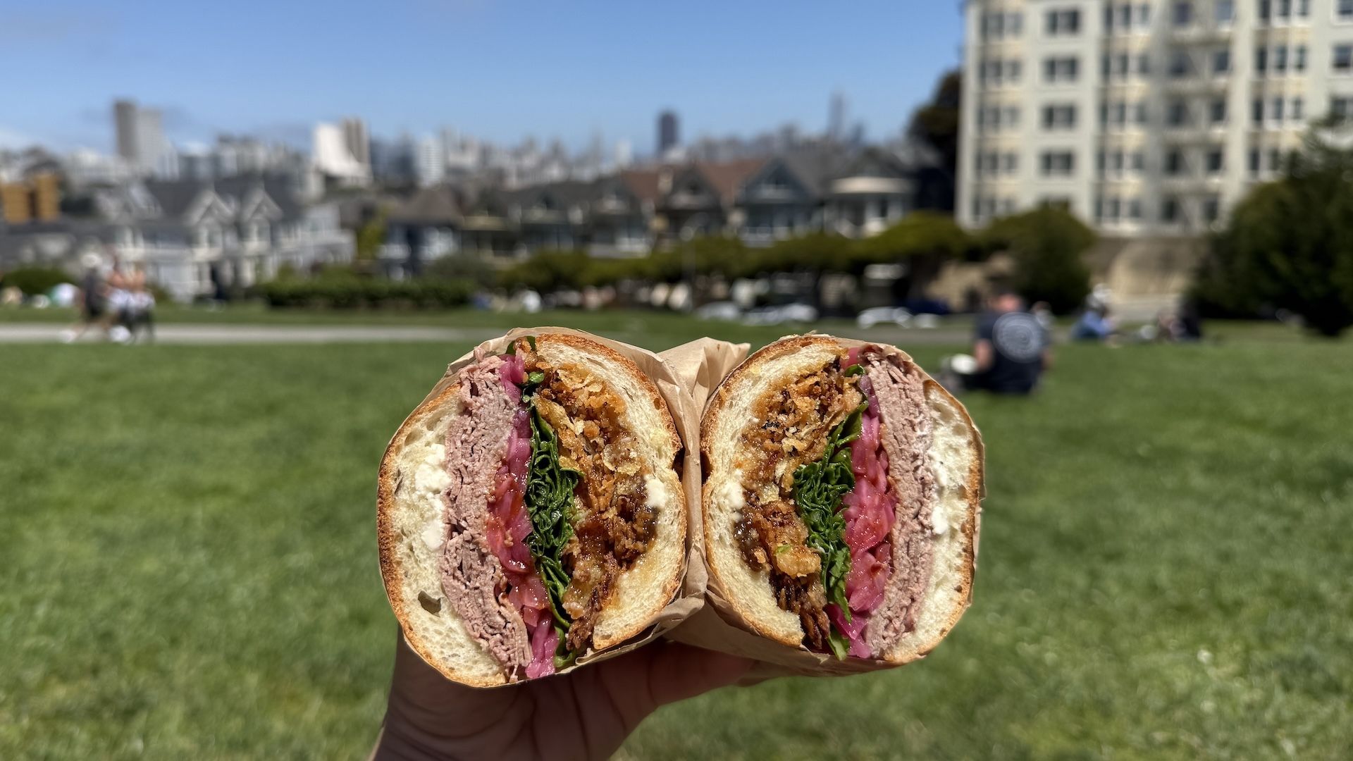 Close-up of a sandwich cut in half showing layers of roast beef, spinach, pickled onions, and crispy fried onions on white bread, held in hand with a park and cityscape in background.
