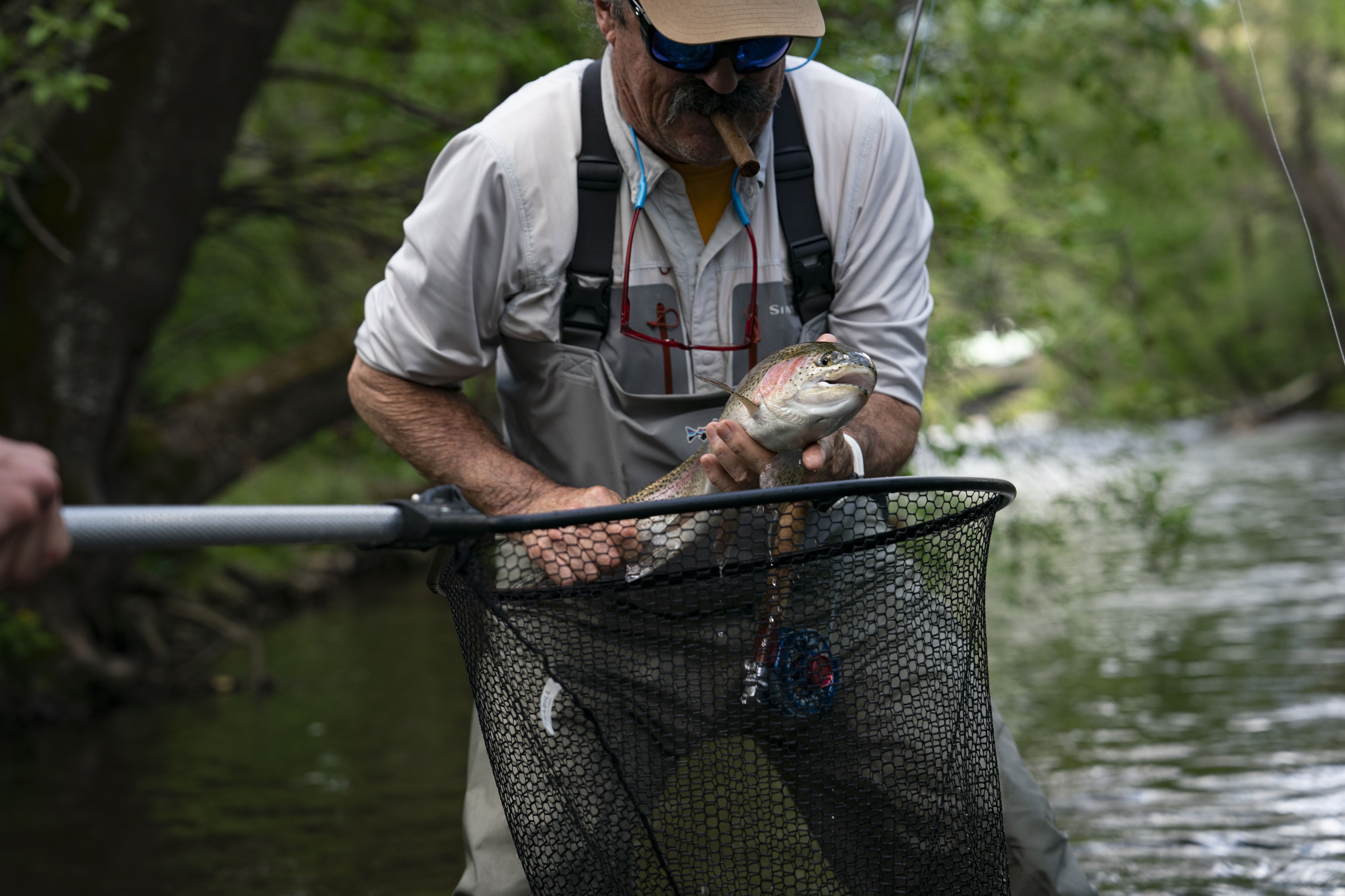 An angler wearing sunglasses, a cap, and waders holds a large rainbow trout above a fishing net while standing in a river.