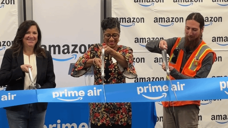 Three people (two Amazon employees and KCK Mayor Christal Watson) holding large white scissors cut a ribbon that says "Prime Air" in front on a backdrop saying "Amazon."
