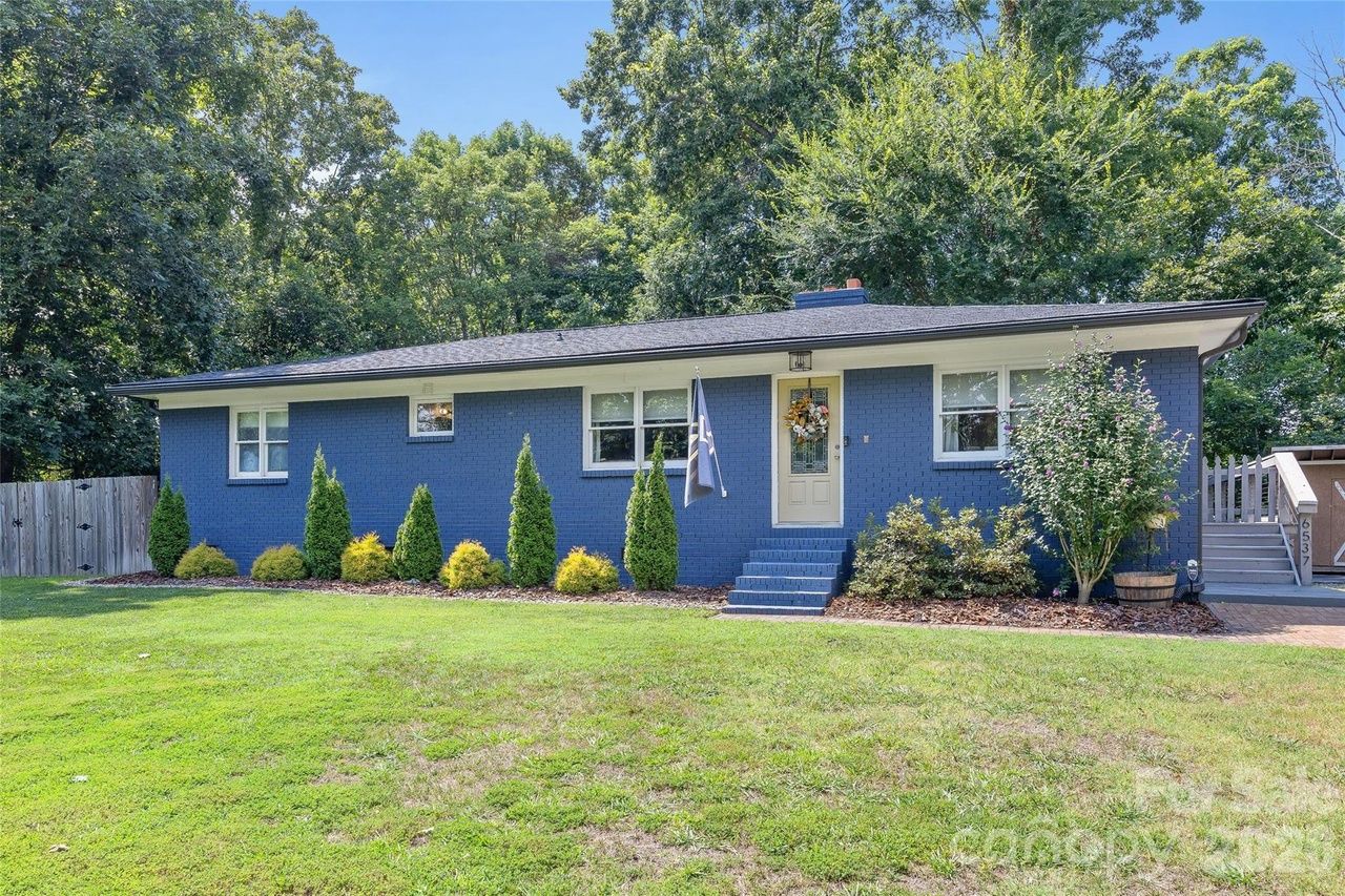 Single-story blue house with white trim sits on a green lawn. Front door with a wreath, blue steps, and a flagpole. Tall evergreens and shrubs line the front bed; fence and trees in the background.