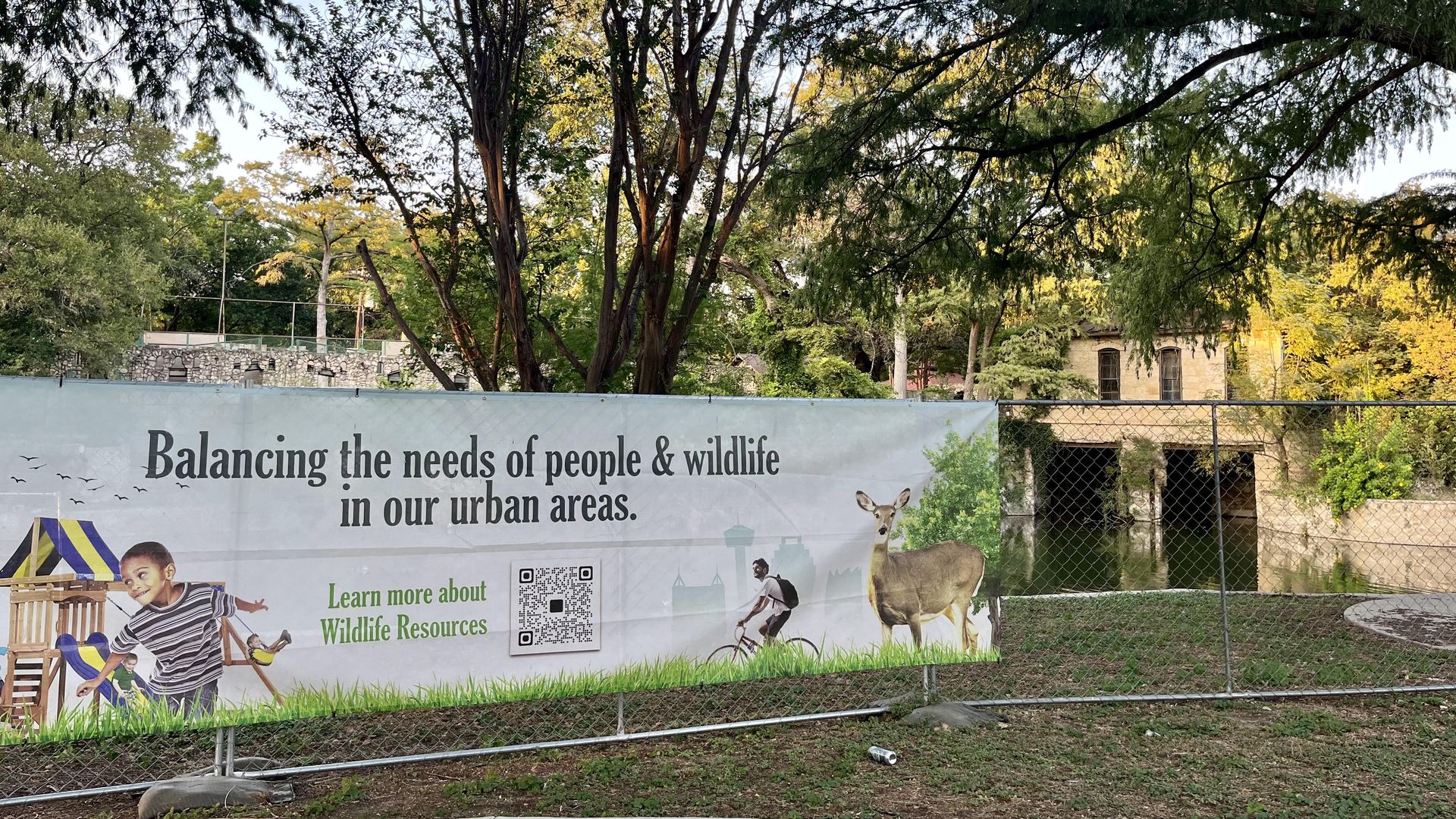 A sign reads "Balancing the needs of people and wildlife in our urban areas," in front of the Pump House and the San Antonio River in Brackenridge Park.