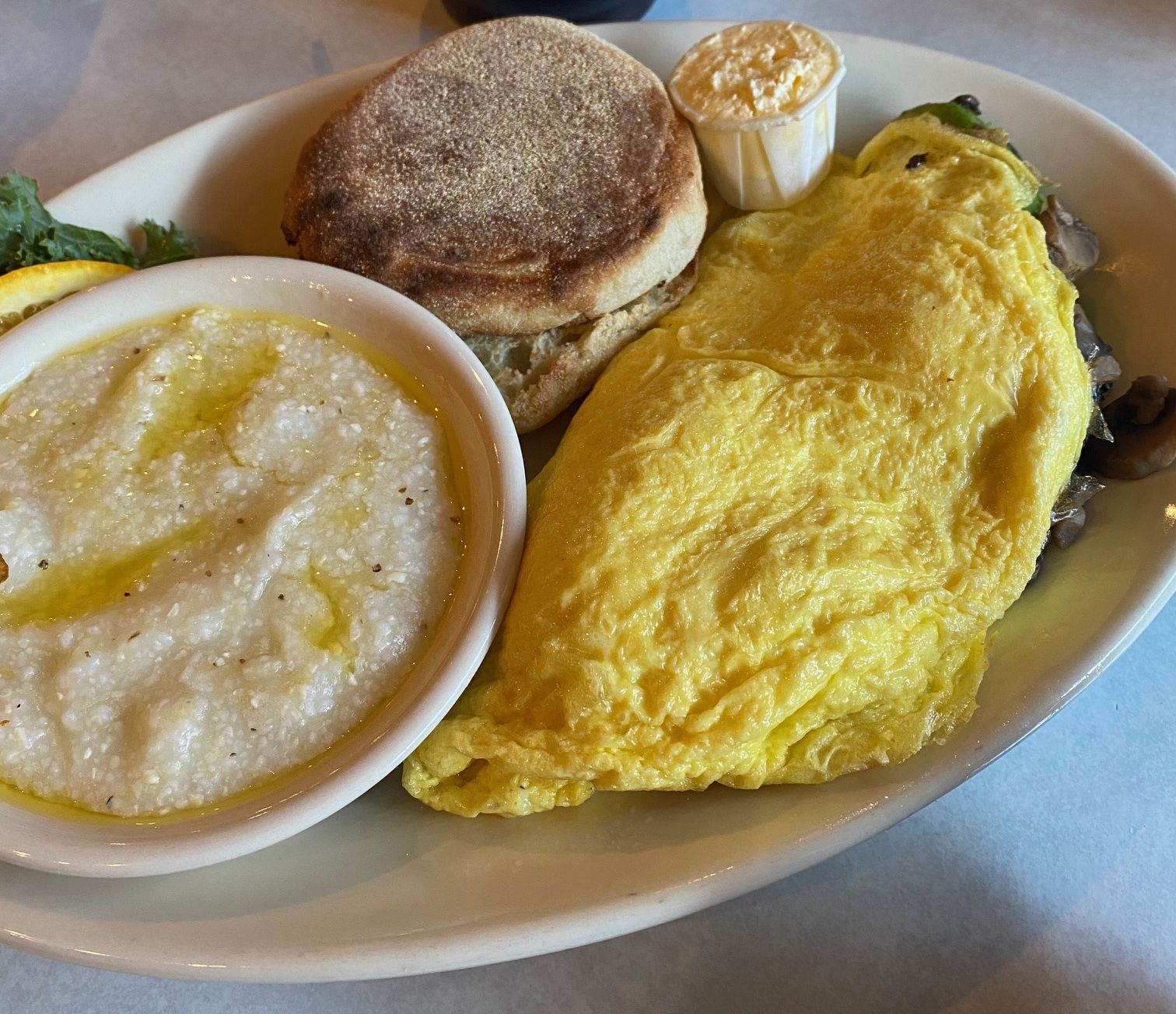 Plate with a yellow folded omelette, an English muffin with butter, and a bowl of creamy grits drizzled with oil and black pepper, garnished with a lemon slice and greens.