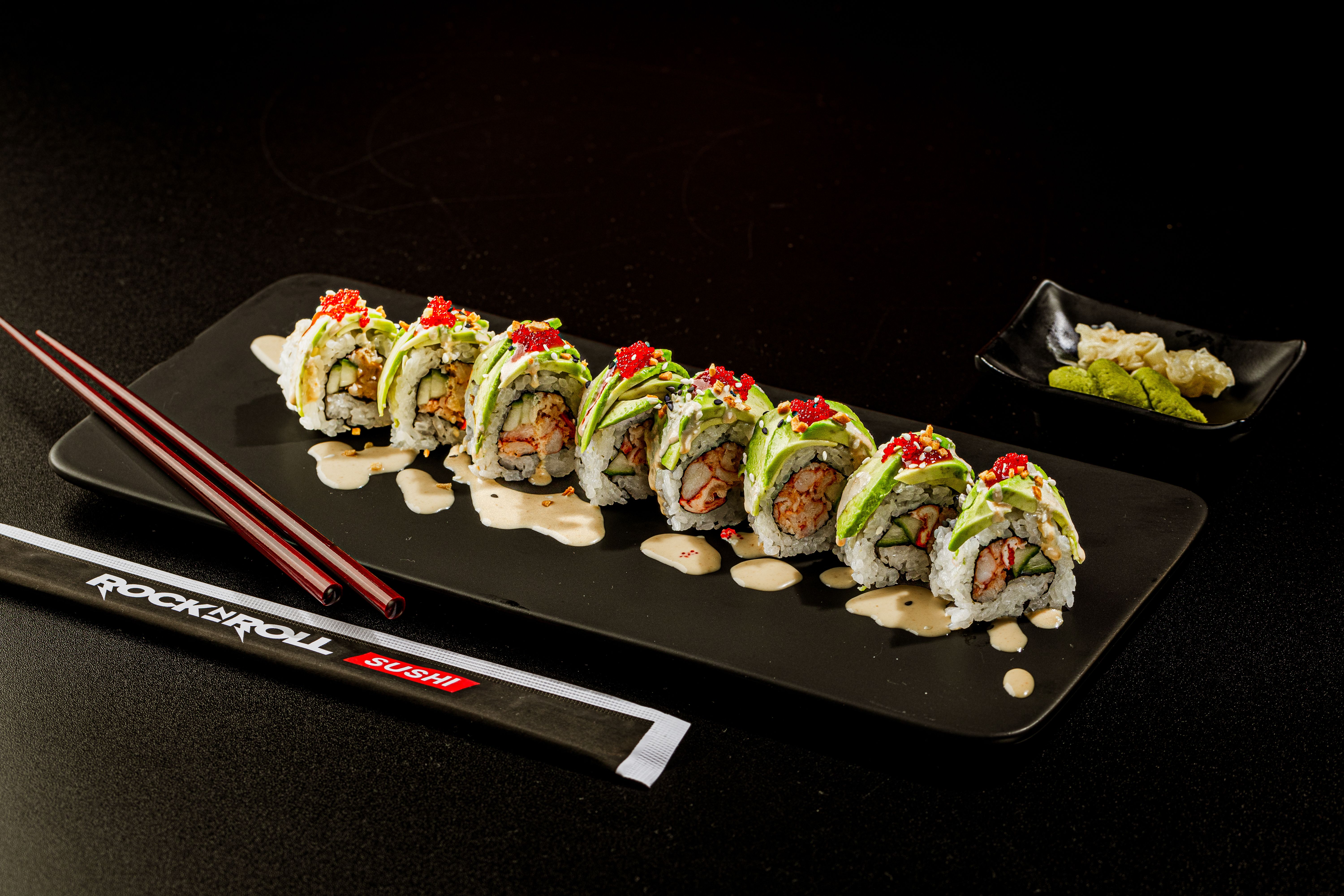 Row of sushi rolls on a black plate, topped with avocado slices and red tobiko, drizzled with sauce. Red chopsticks rest nearby; a small plate of wasabi and pickled ginger sits in the background.