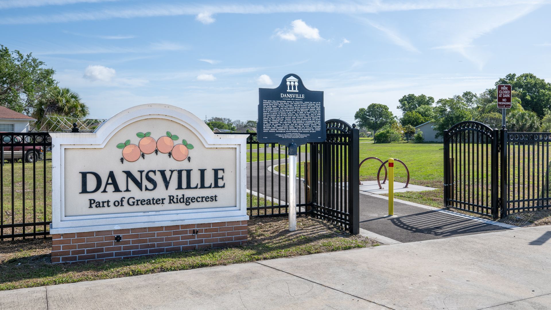 Entrance to Dansville: a white sign with "DANSVILLE" and "Part of Greater Ridgecrest" below orange icons on a brick base, beside black iron gates leading to a green park under a blue sky.