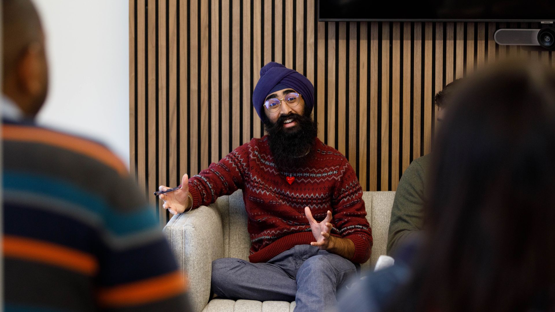 A bearded man wearing a purple turban and glasses sits on a beige sofa in a meeting room, wearing a red knitted sweater. He gestures with his hands as others listen.