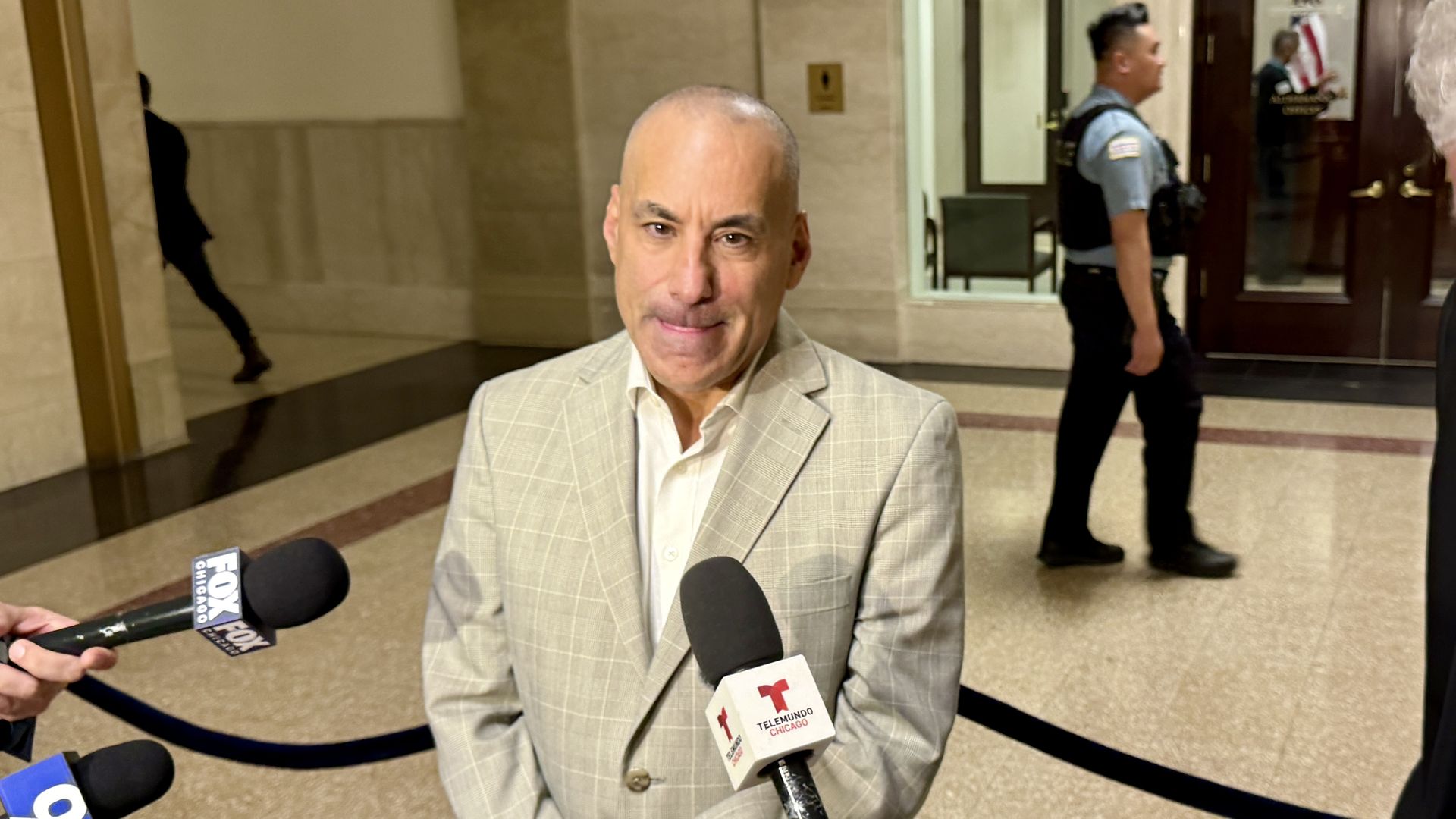 Bald man in a light gray plaid suit is interviewed by reporters in a marble lobby; microphones include Fox Chicago and Telemundo as a security officer stands by a door with an American flag.