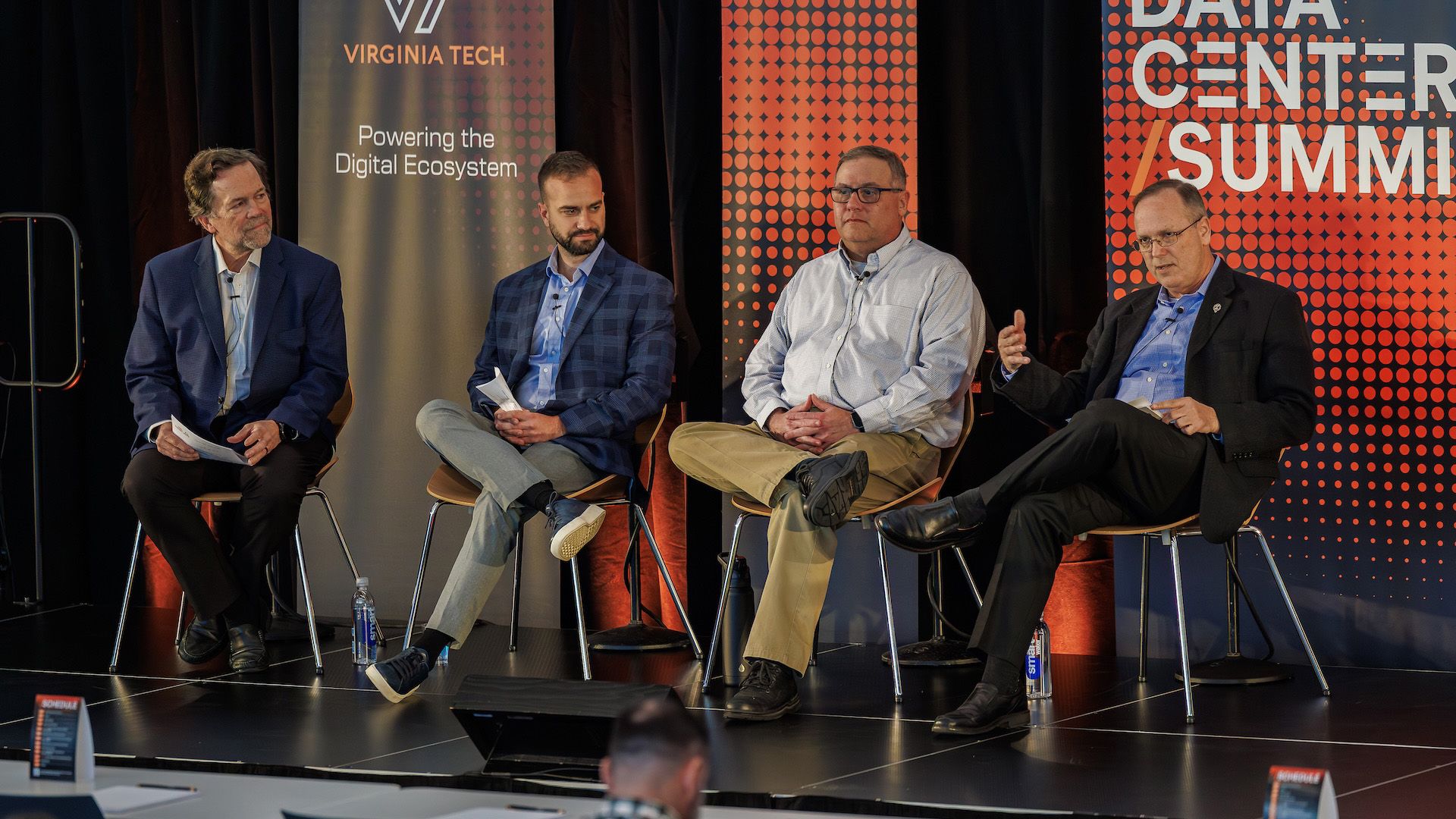 Four men sit on a panel stage at a conference, with Virginia Tech banners reading "Powering the Digital Ecosystem" and "Data Center Summit." They wear blazers and clip-on mics.