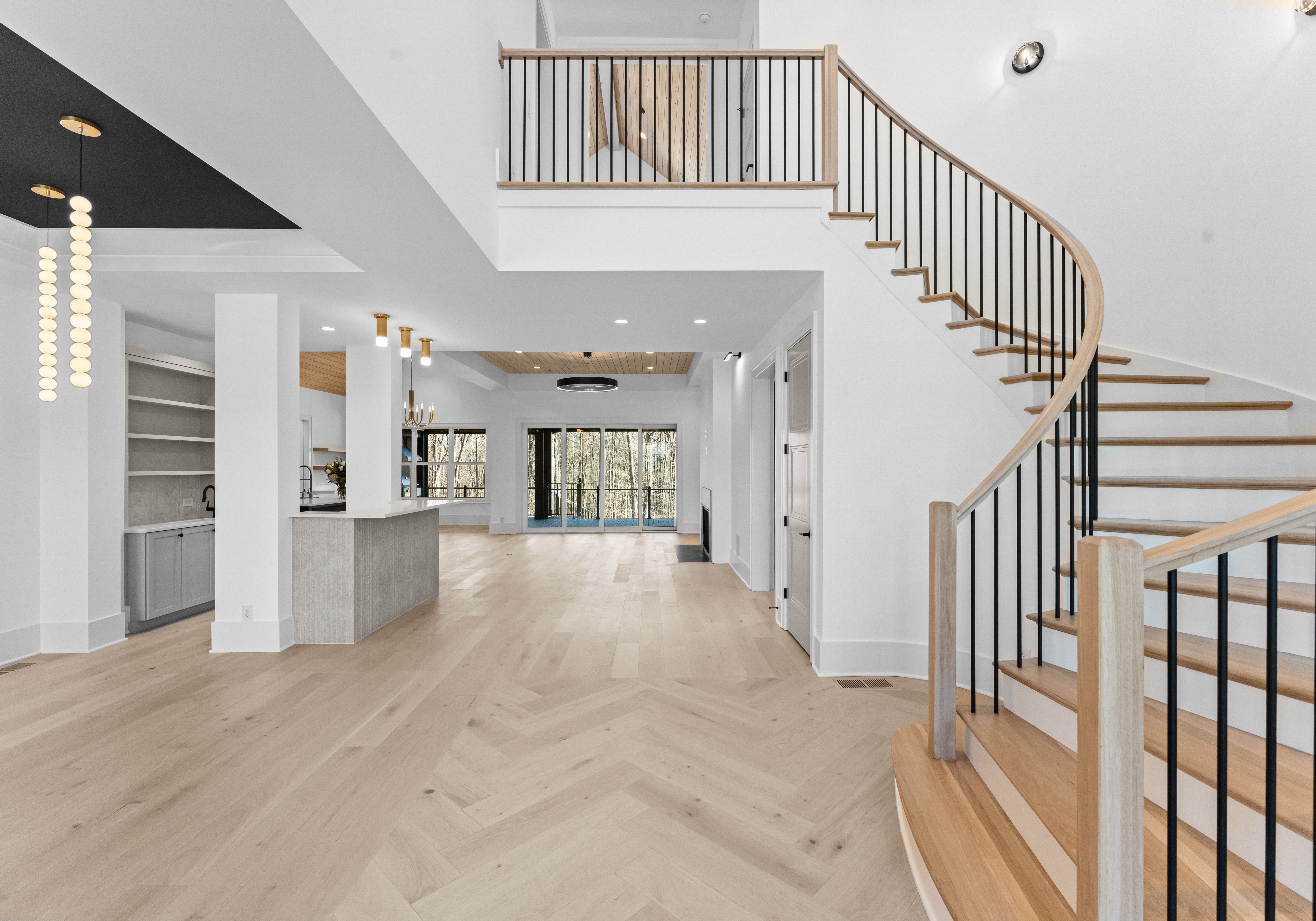 Spacious modern open-plan interior with light wood flooring, white walls, curved wood staircase with black metal railings, hanging pendant lights, kitchen island, large windows overlooking trees.