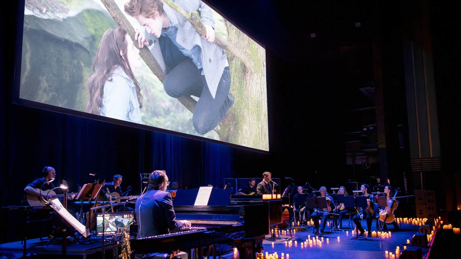A music ensemble plays on a stage while Twlight plays on the screen