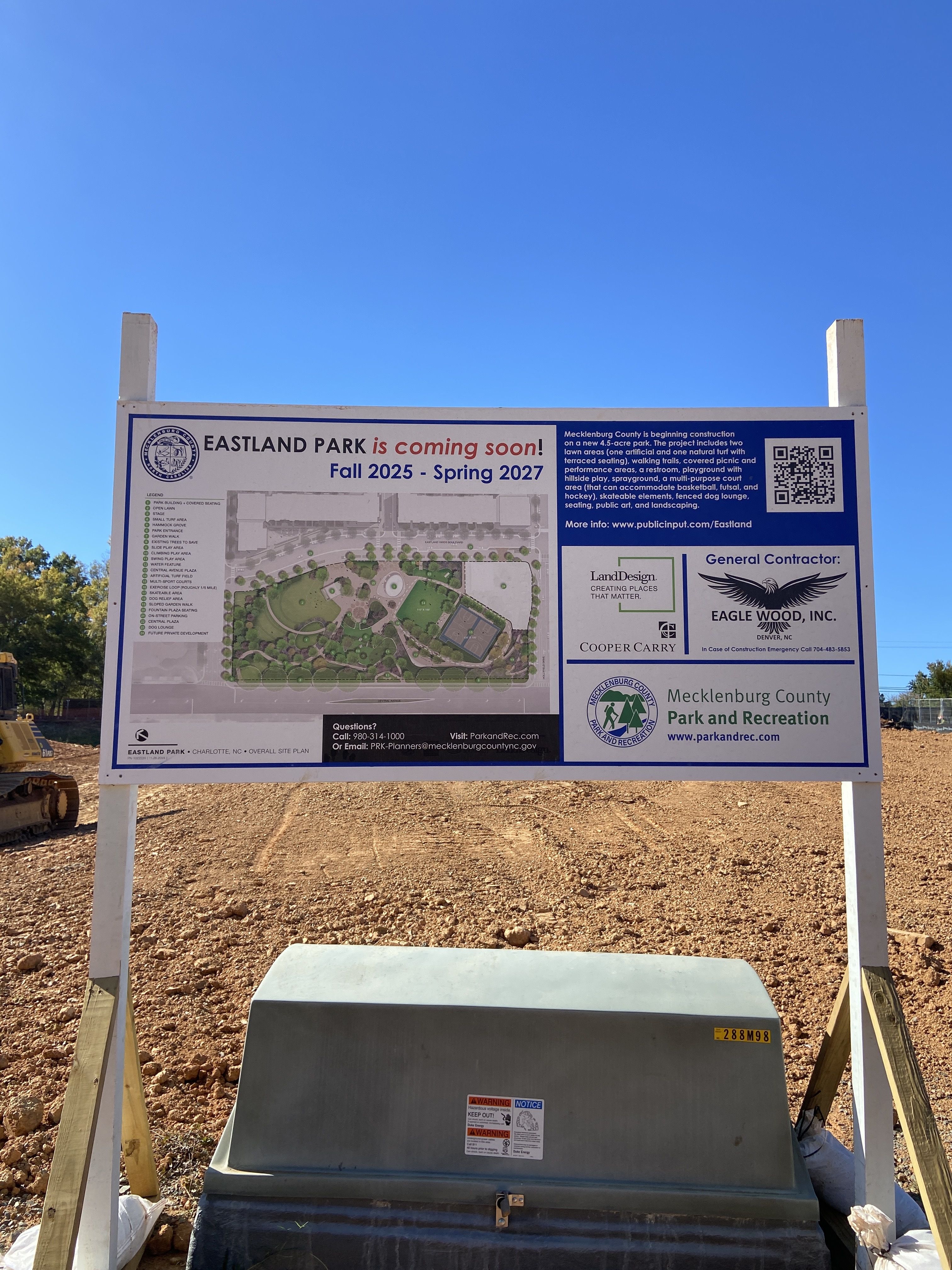 Construction site sign for Eastland Park coming Fall 2025 to Spring 2027 in Charlotte, NC, with park map and project details under a clear blue sky.