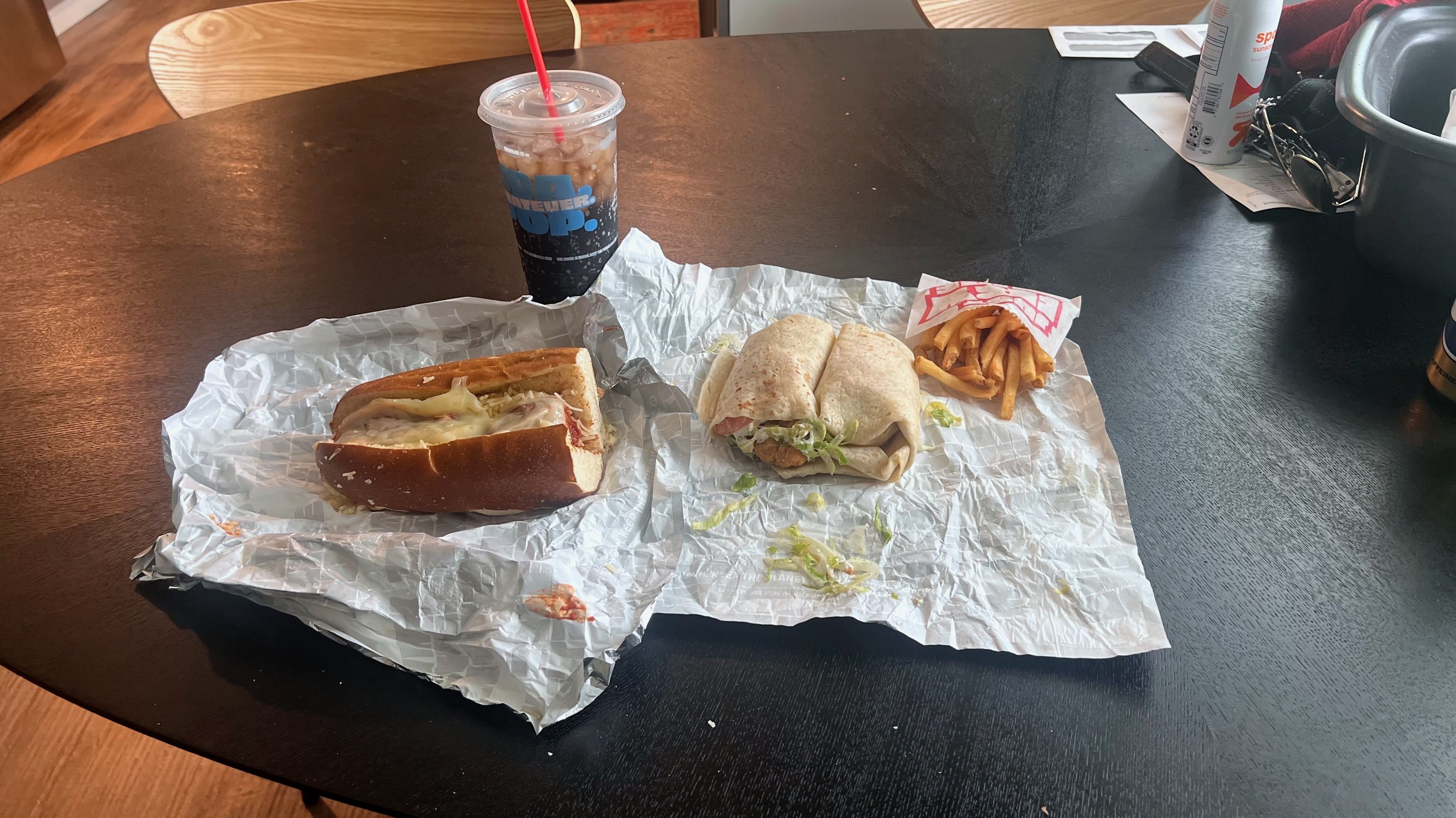 An overview of a drink, meatball sub, chicken wraps and fries on a wrapper on a table