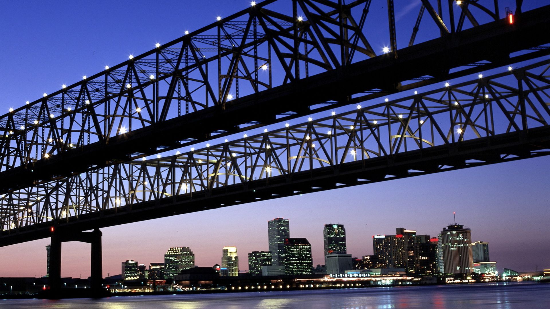 The New Orleans city skyline is viewed from beneath the twin span of the Crescent City Connection at dusk. Lights are seen along the bridge.
