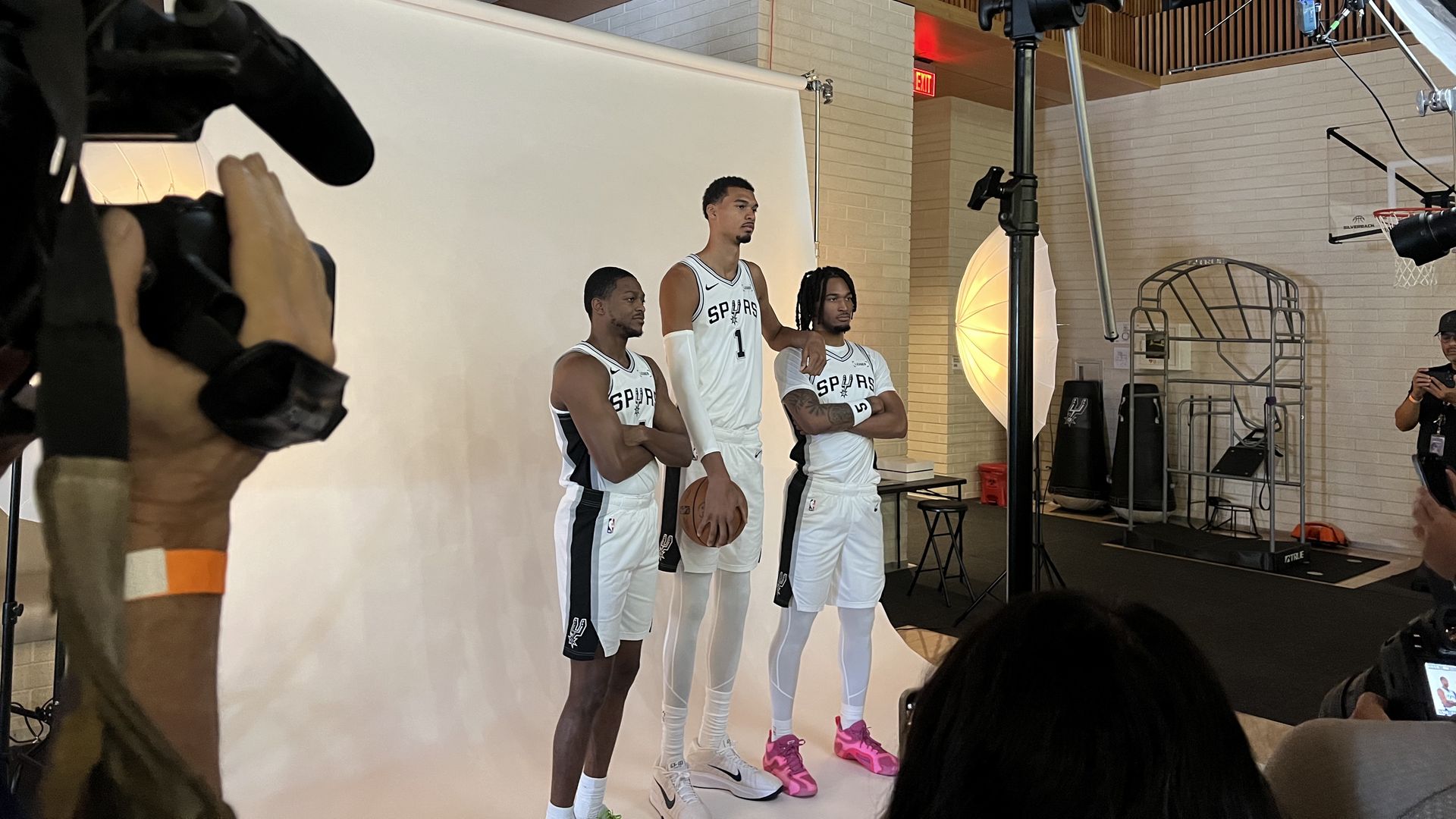 Three San Antonio Spurs basketball players wearing white uniforms pose in front of a white backdrop during a photo shoot with cameras and lighting equipment visible.