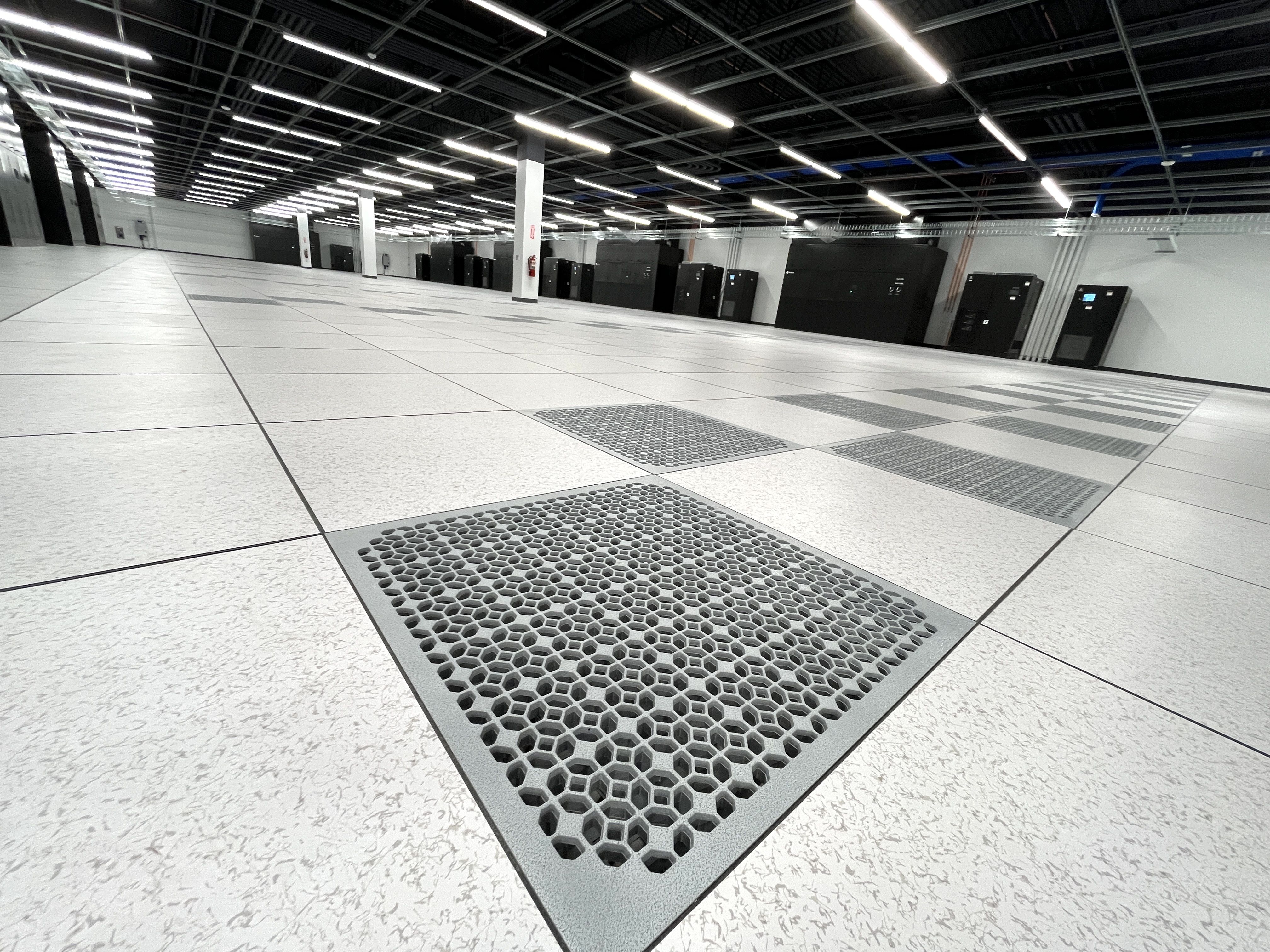 Wide view of a modern data center room with clean white floor tiles, hexagonal ventilation grates, bright overhead lights, and black server cabinets along the walls.