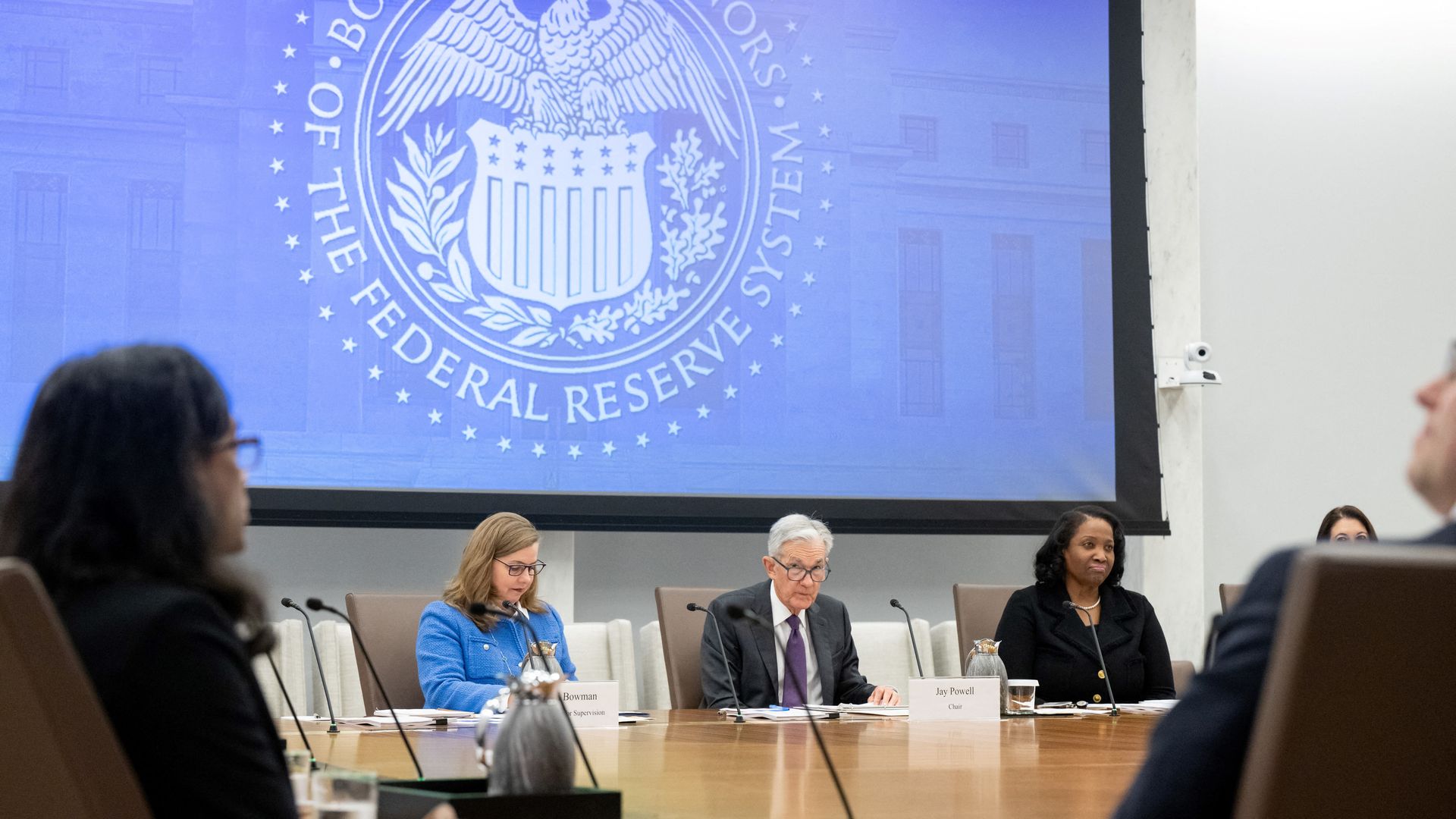 Federal Reserve Board of Governors meeting with members seated at a large table. A blue screen with the Board's eagle seal logo is projected behind them.