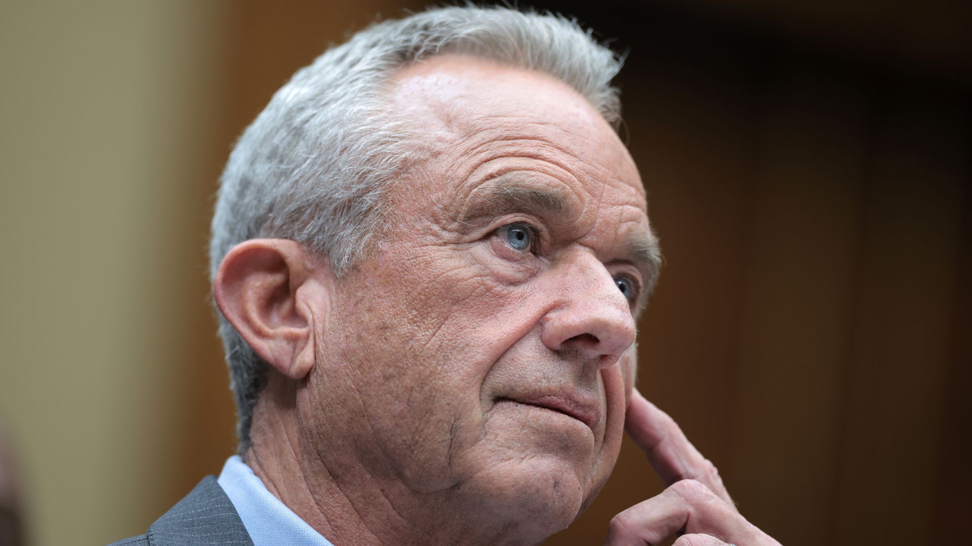 Close-up of an older man with gray hair and blue eyes wearing a gray suit and light blue shirt, resting his finger on his cheek in a thoughtful pose.