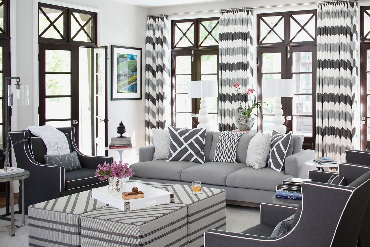 Bright living room with gray and white chevron curtains, gray sofa with patterned pillows, dark armchairs, striped ottoman coffee table, and white lamps by large wooden windows.