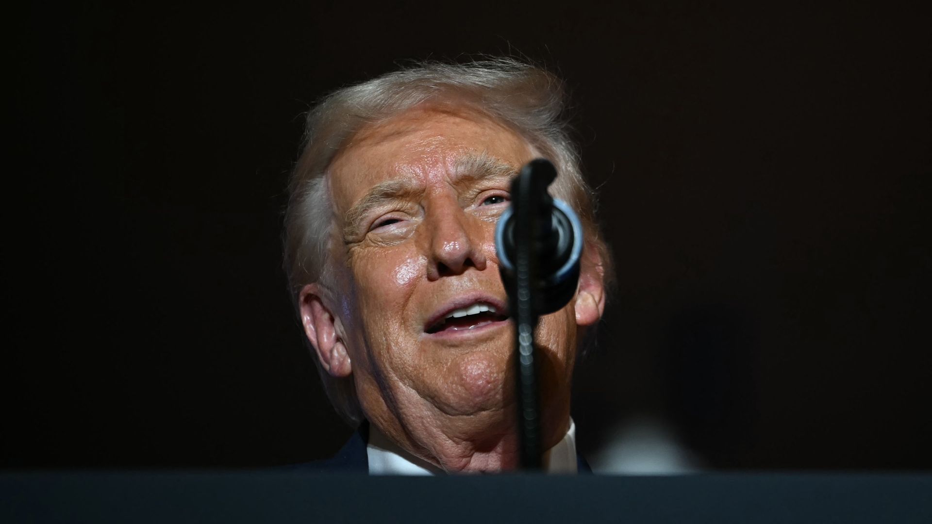 Close-up of a man with light hair speaking into a microphone against a dark background, his face illuminated and showing an expressive look.