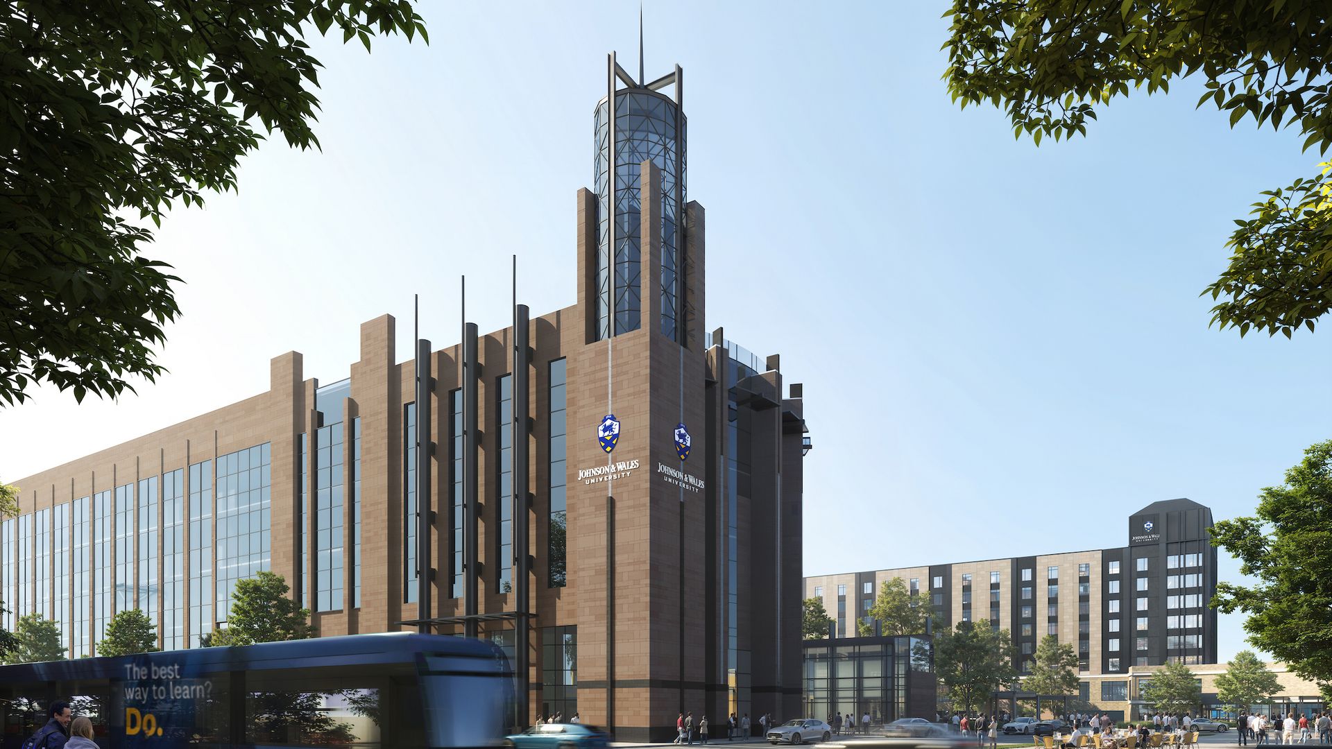 A modern brick and glass building with a cylindrical glass tower and metal spires rises above a street. JWU logos on the facade, trees framing the scene, a blue bus and pedestrians nearby.