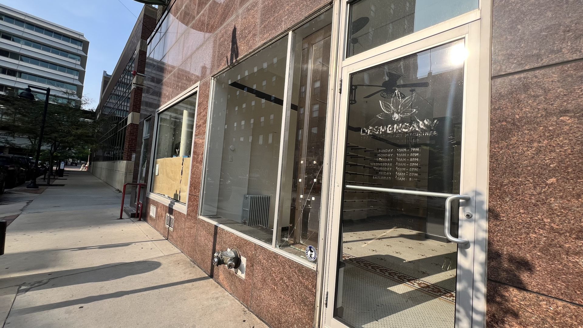 Street view of a building with brown marble walls and large windows, one partly covered with paper. A glass door with store hours and a leaf logo is visible, reflecting sunlight.