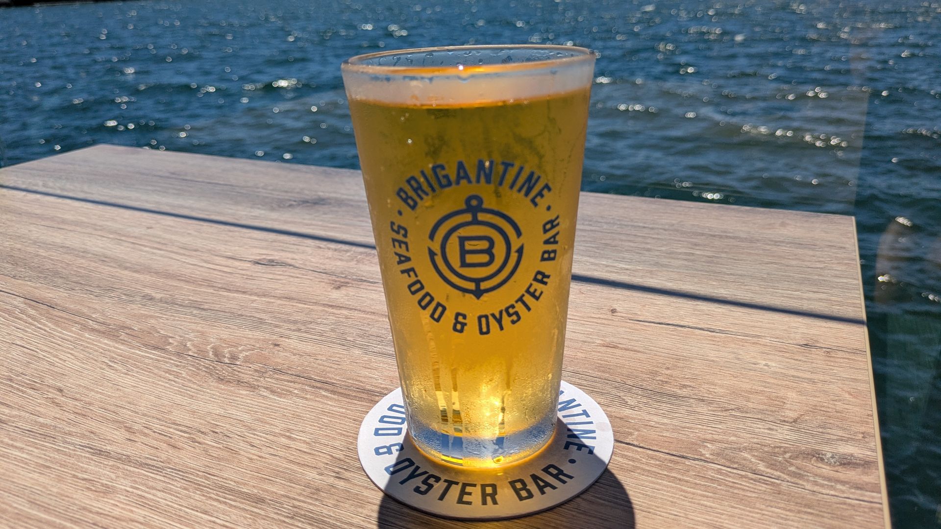Beer glass with Brigantine logo on a wooden table, condensation beads; blue harbor water, distant shoreline and small boats under a sunny sky.