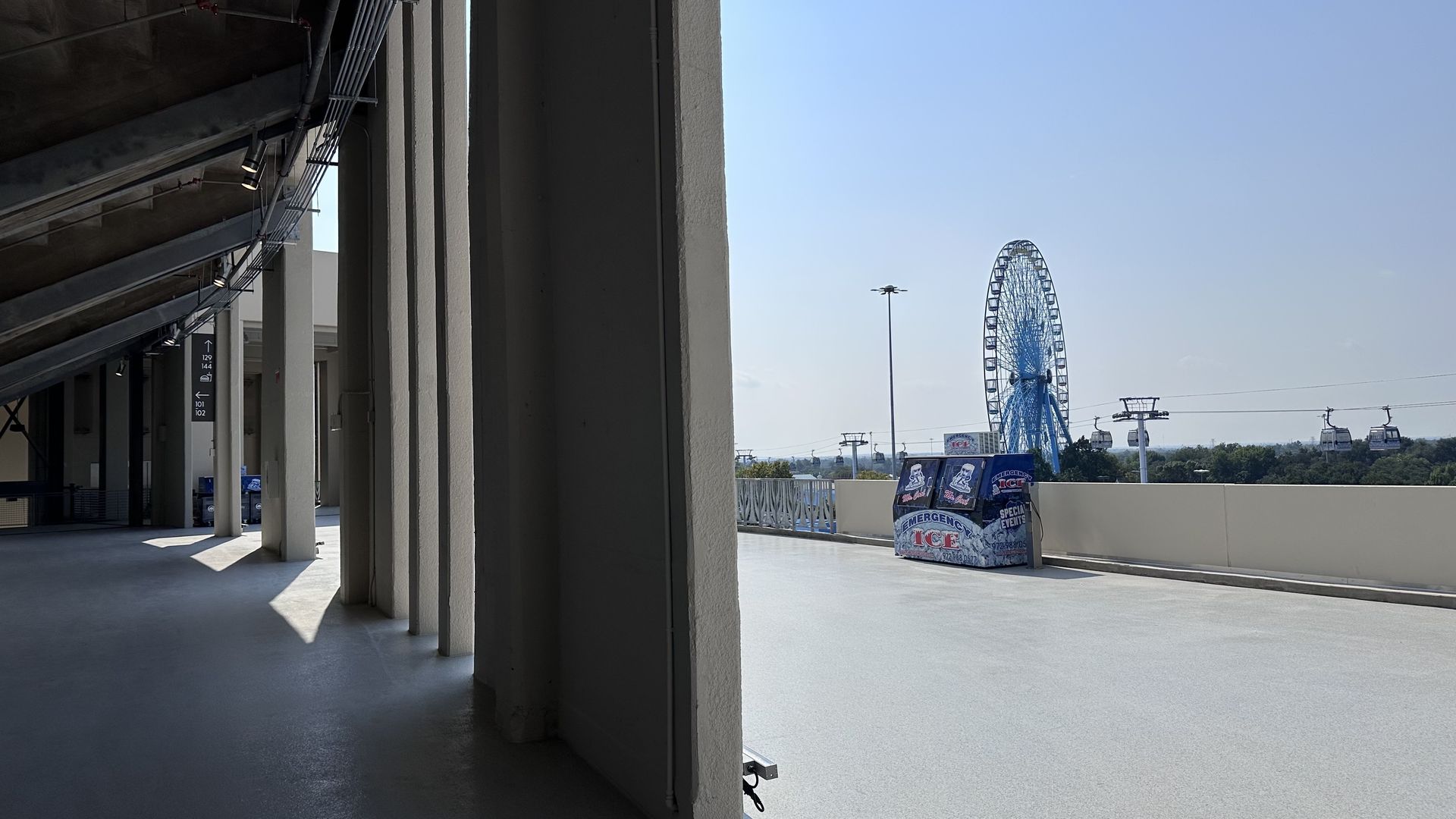 Part of a shaded building with tall white columns on the left contrasts with a bright open area on the right featuring a blue Ferris wheel, cable cars, and an Emergency Ice box.