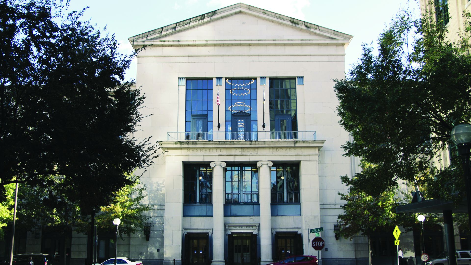 Front view of the white stone Nashville Public Library building with tall columns and large blue-tinted windows, trees on both sides, and cars parked on the street.