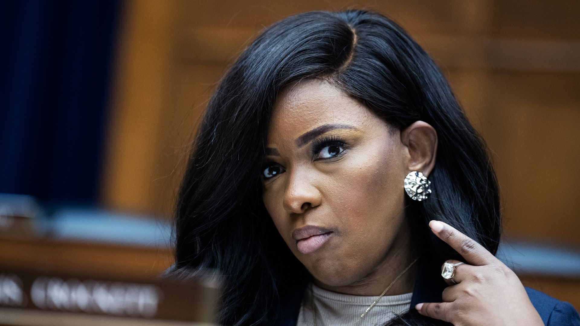 Jasmine Crockett, wearing a blue suit and sitting in a wooden room in front of a blurry placard with her name on it.