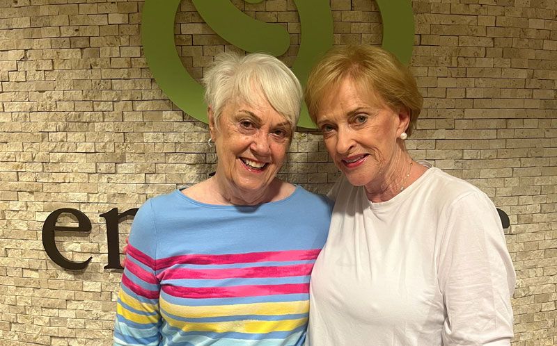 Two women smiling warmly while standing closely together against a textured stone wall backdrop.